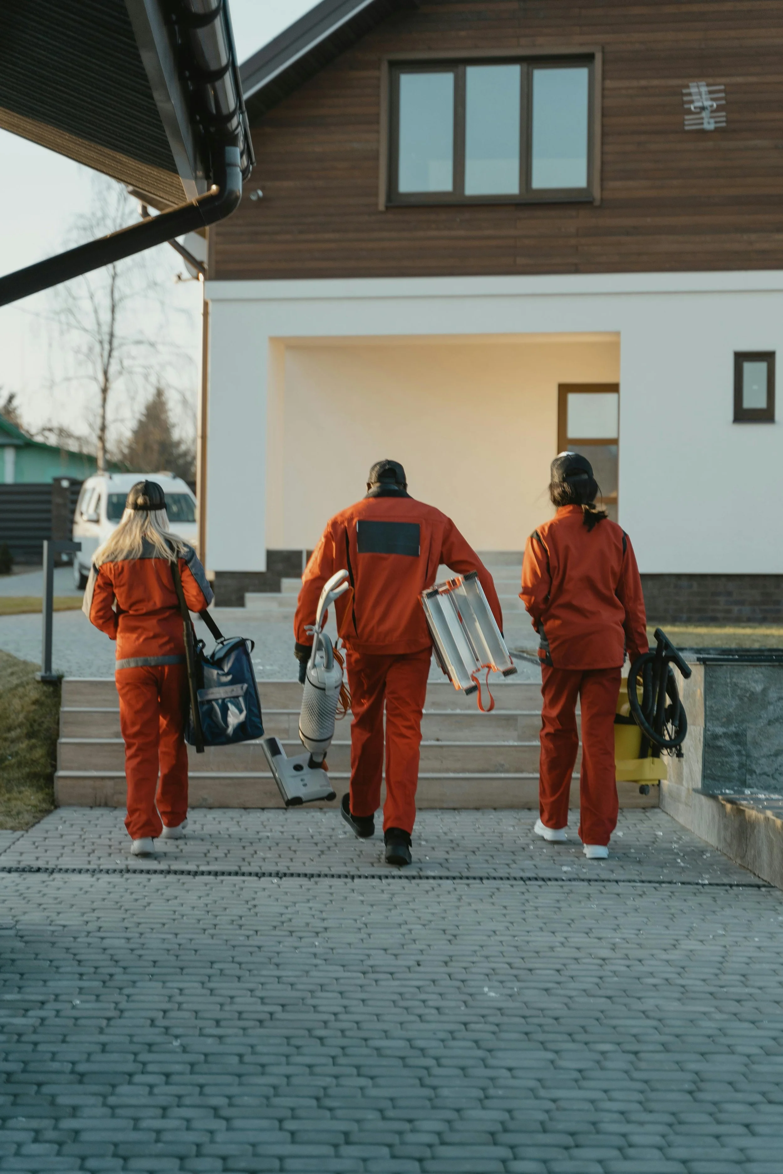 Three deep cleaners in uniforms walking up the front steps of a modern house, carrying cleaning equipment.