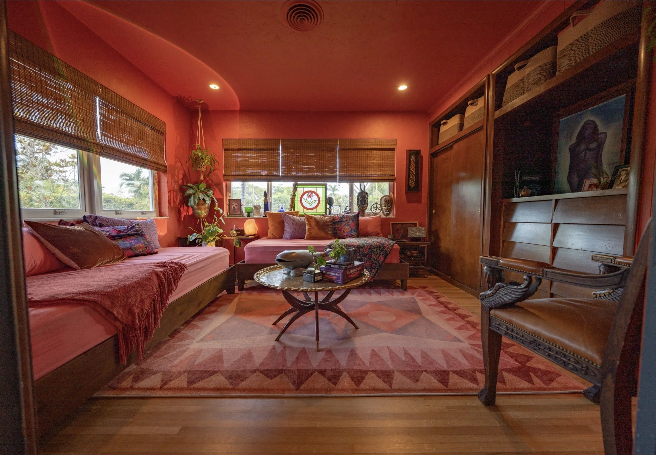 Cozy living room with pink and purple cushions, a round coffee table with decorative items, and a window with bamboo blinds. Decor includes sculptures, plants, and colorful wall art.