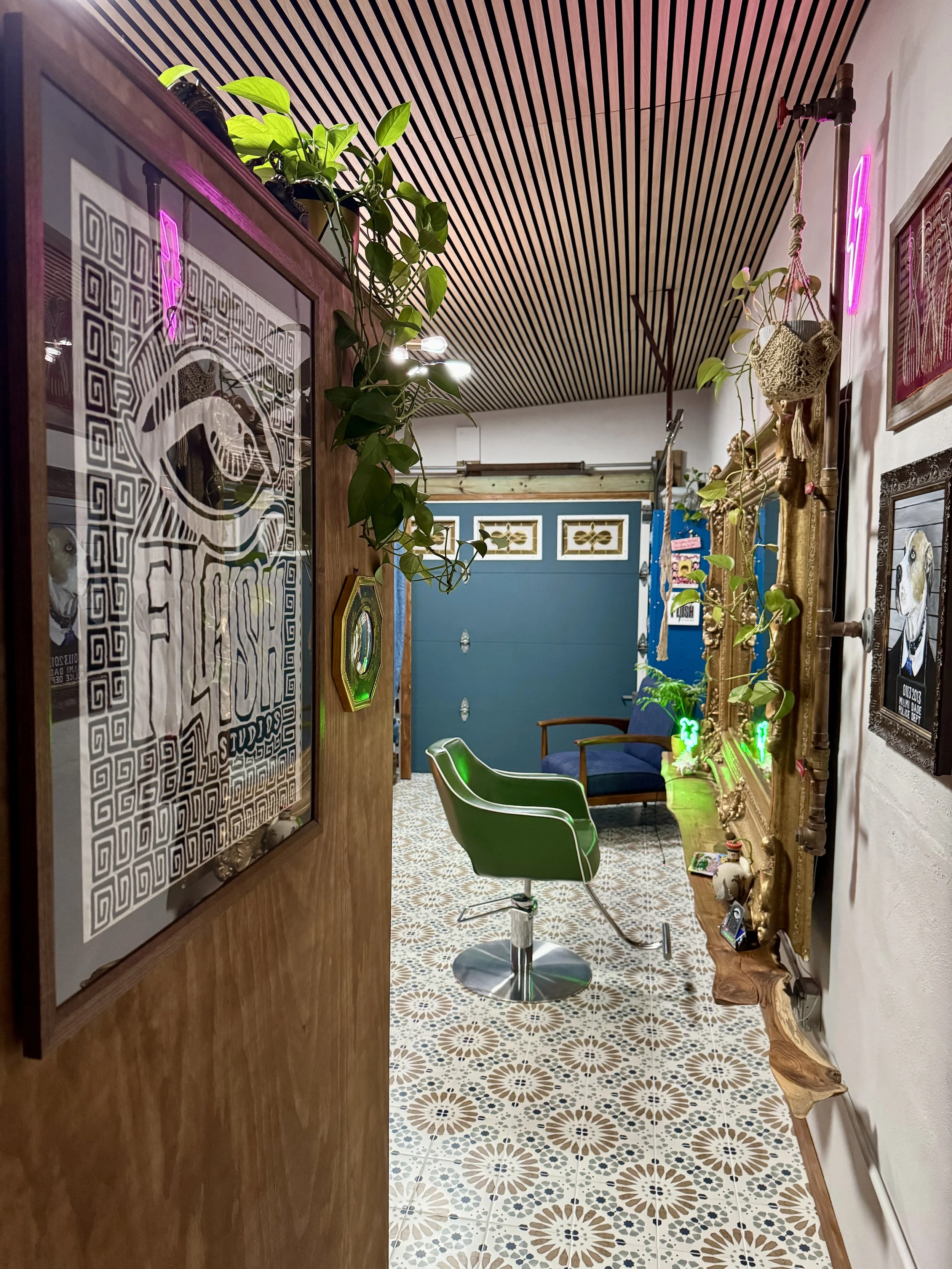Interior view of a cozy room with a patterned tile floor, a wooden door with artwork and plants, a green salon chair, a blue chair, decorative wall art, neon lights, and hanging plants.