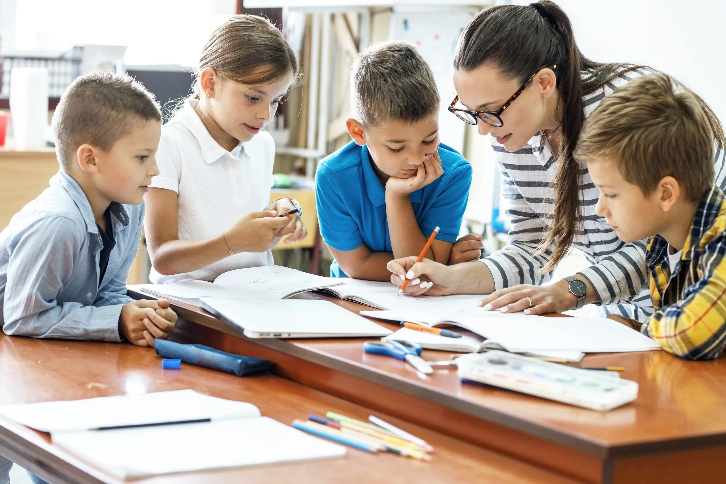 A teacher and four students gathered around a table looking at a workbook, with books and supplies on the table in a classroom.