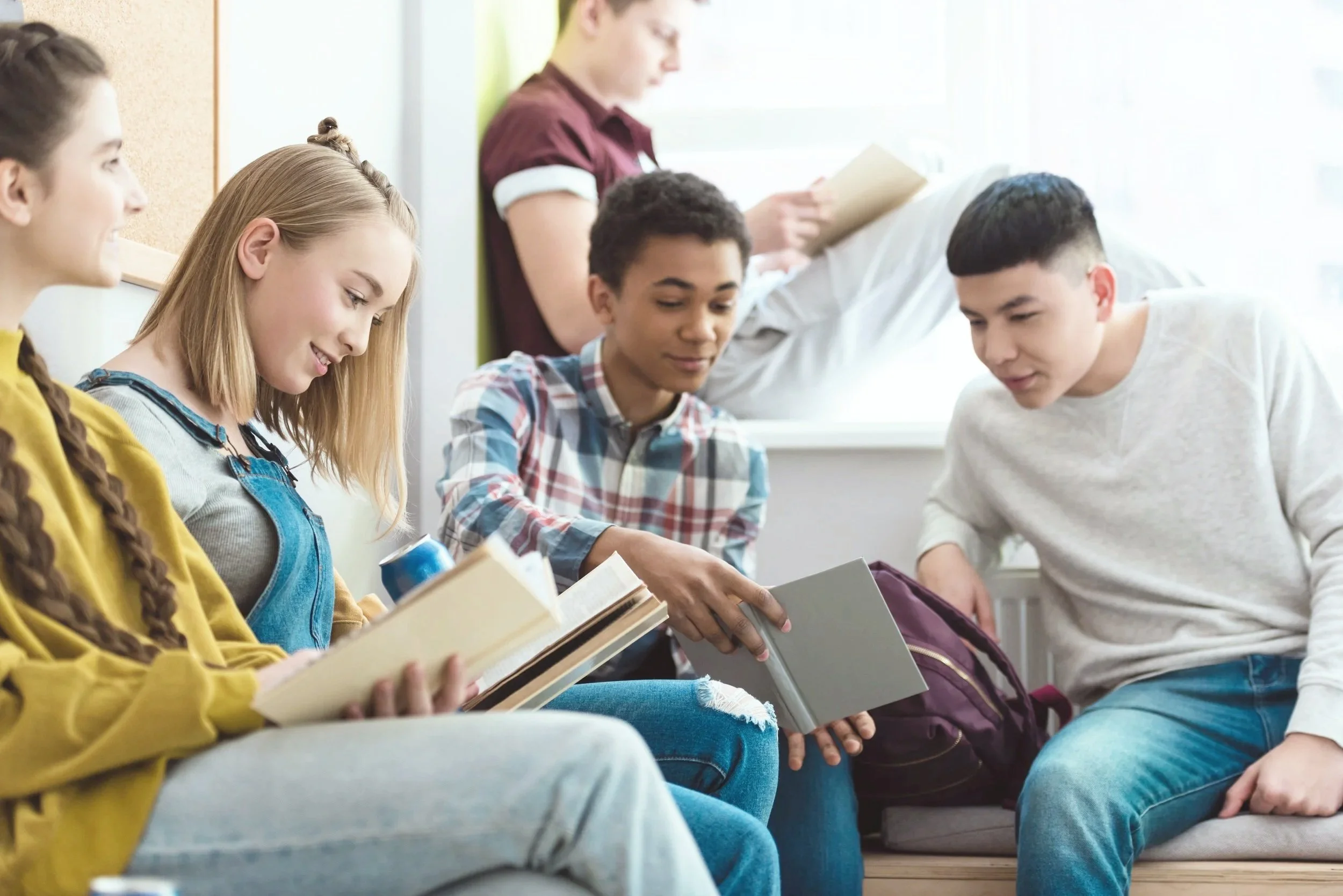 Group of five diverse teenagers sitting and standing in a classroom, engaging with books and notebooks, smiling and talking.