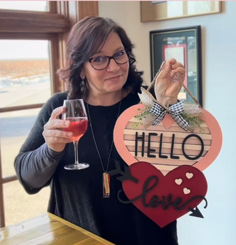 A woman with glasses holding a glass of red wine and a Valentine’s Day sign that says 'Hello Love' with a heart and arrow, inside a room with large windows.