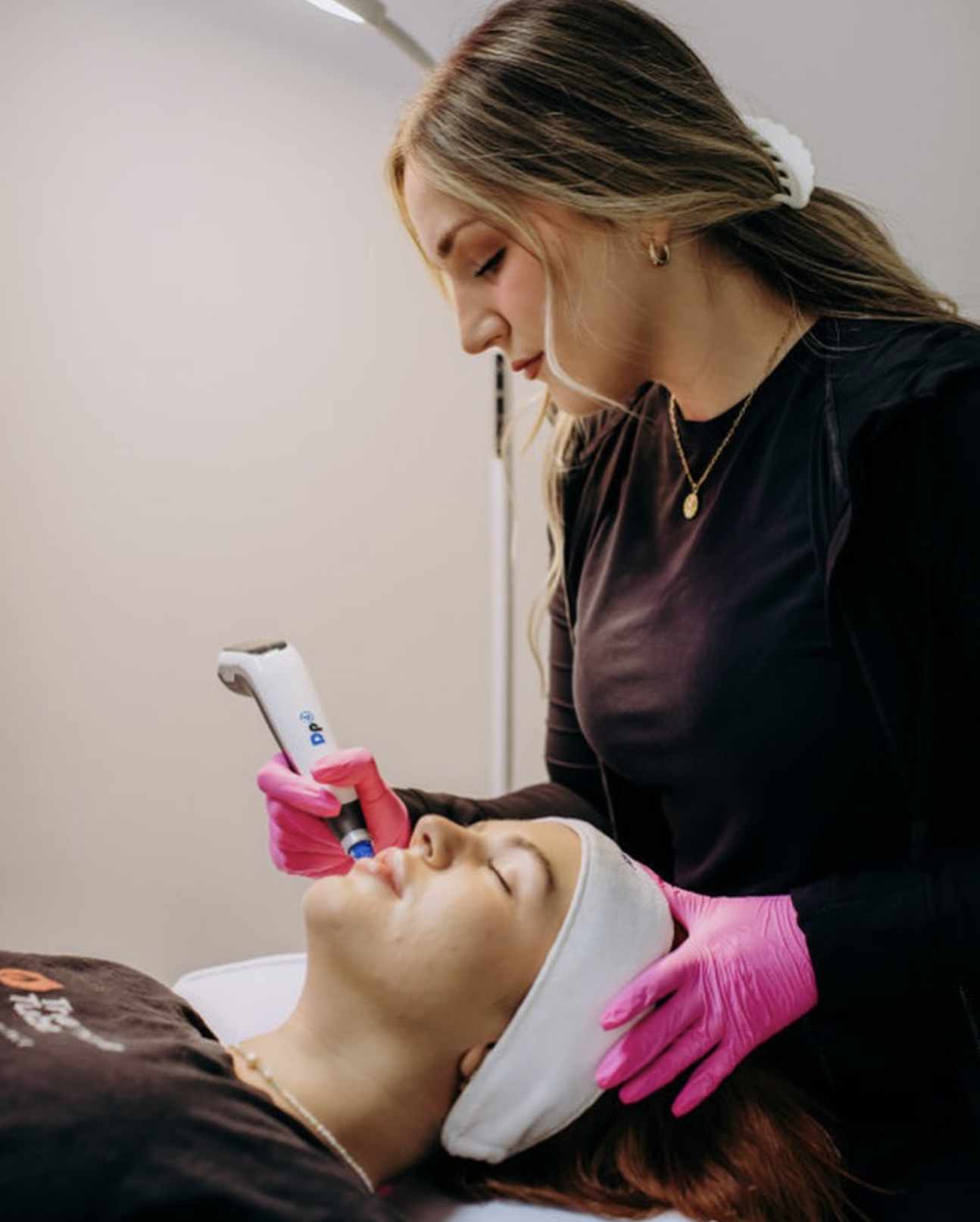 A photo of Emily, from 707 Laser & Esthetics, performing a cosmetic skin treatment on a woman lying down with a headband. The practitioner is wearing pink gloves and holding a skincare device to the patient's face.