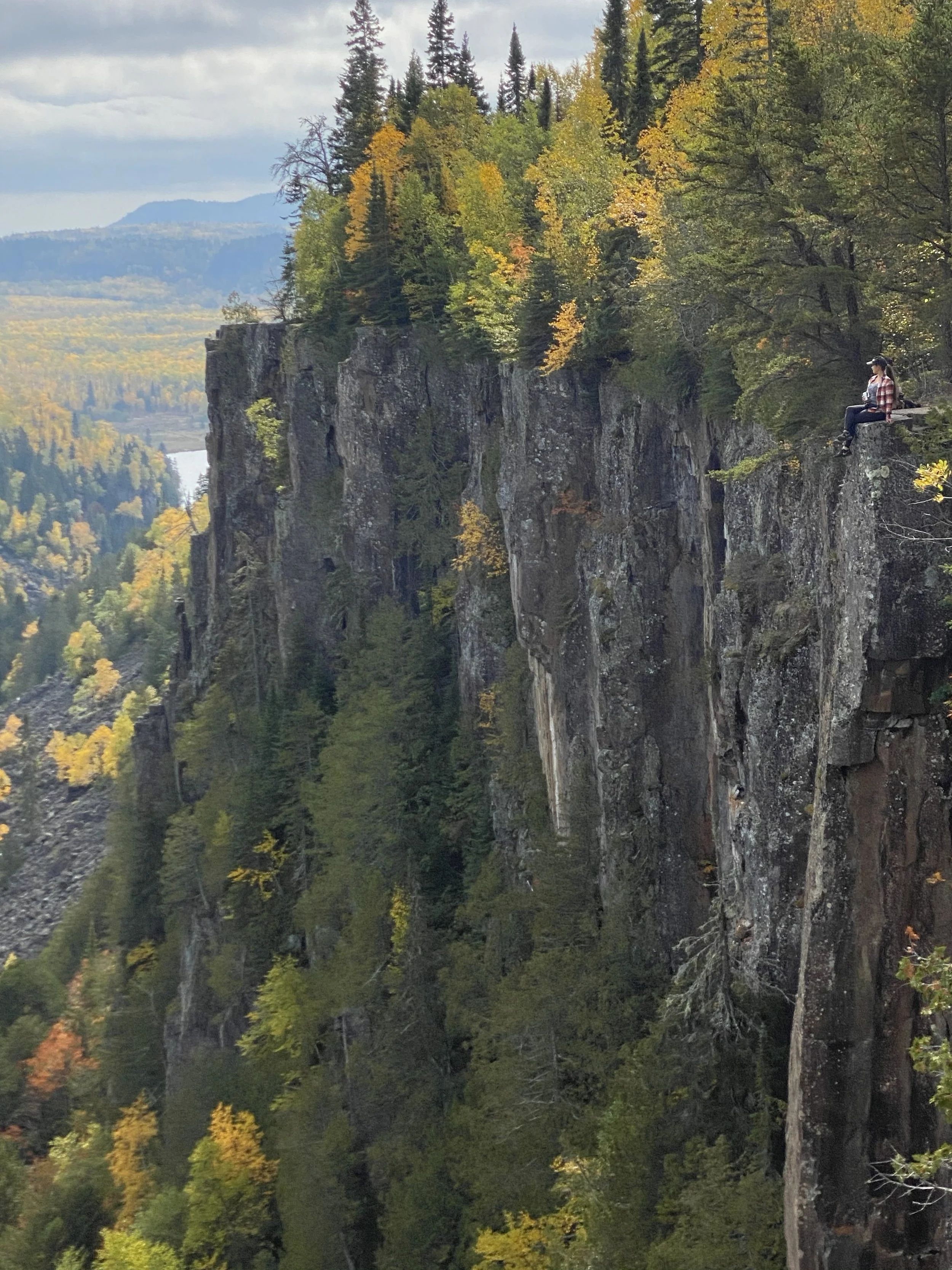 A photo of Julia sitting on the edge of one of the many tall cliffs at Ouimet canyon near Thunder Bay surrounded by trees and fall colours.