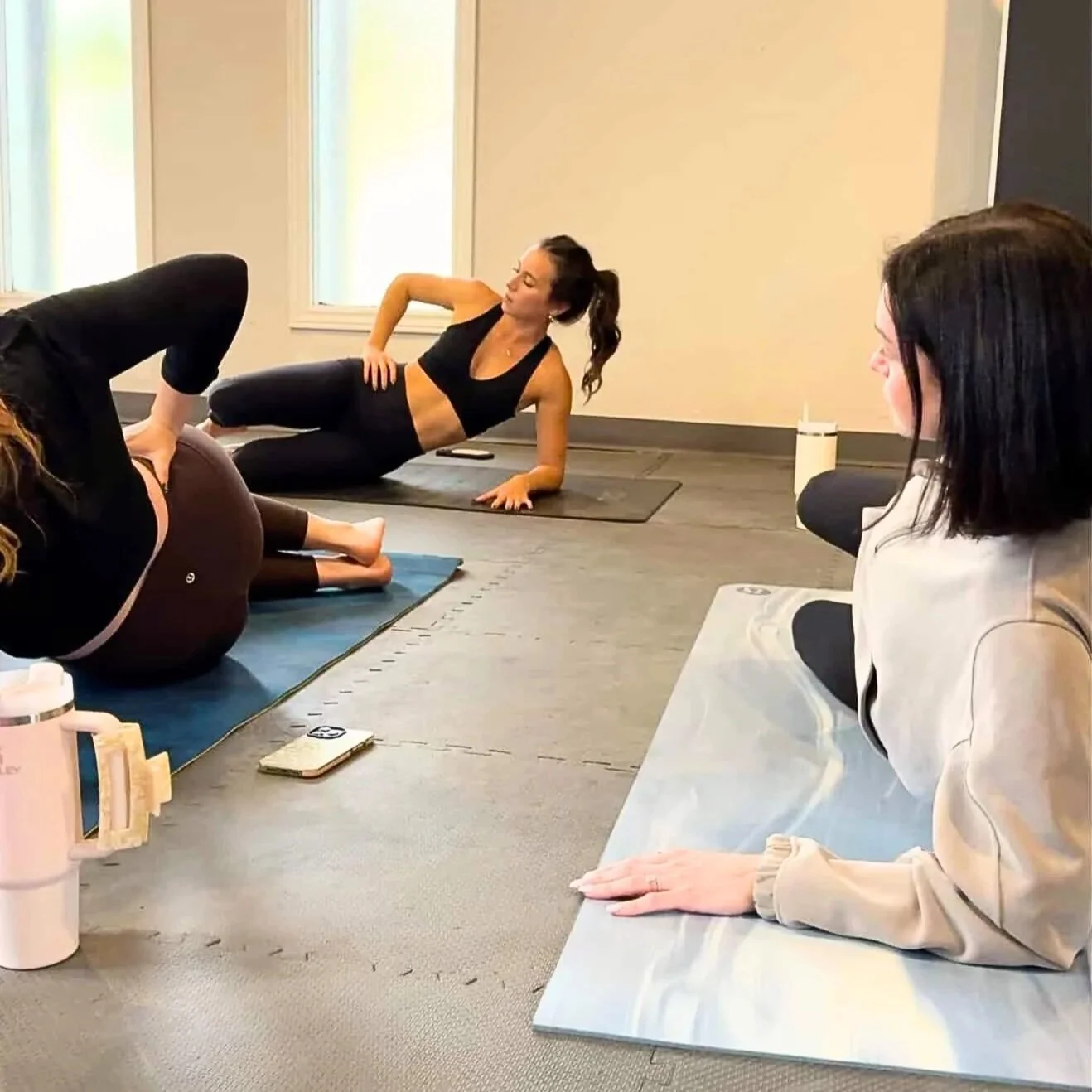 A photo of Sarina leading her pilates class with a group of women participating on mats in a bright room with large windows.