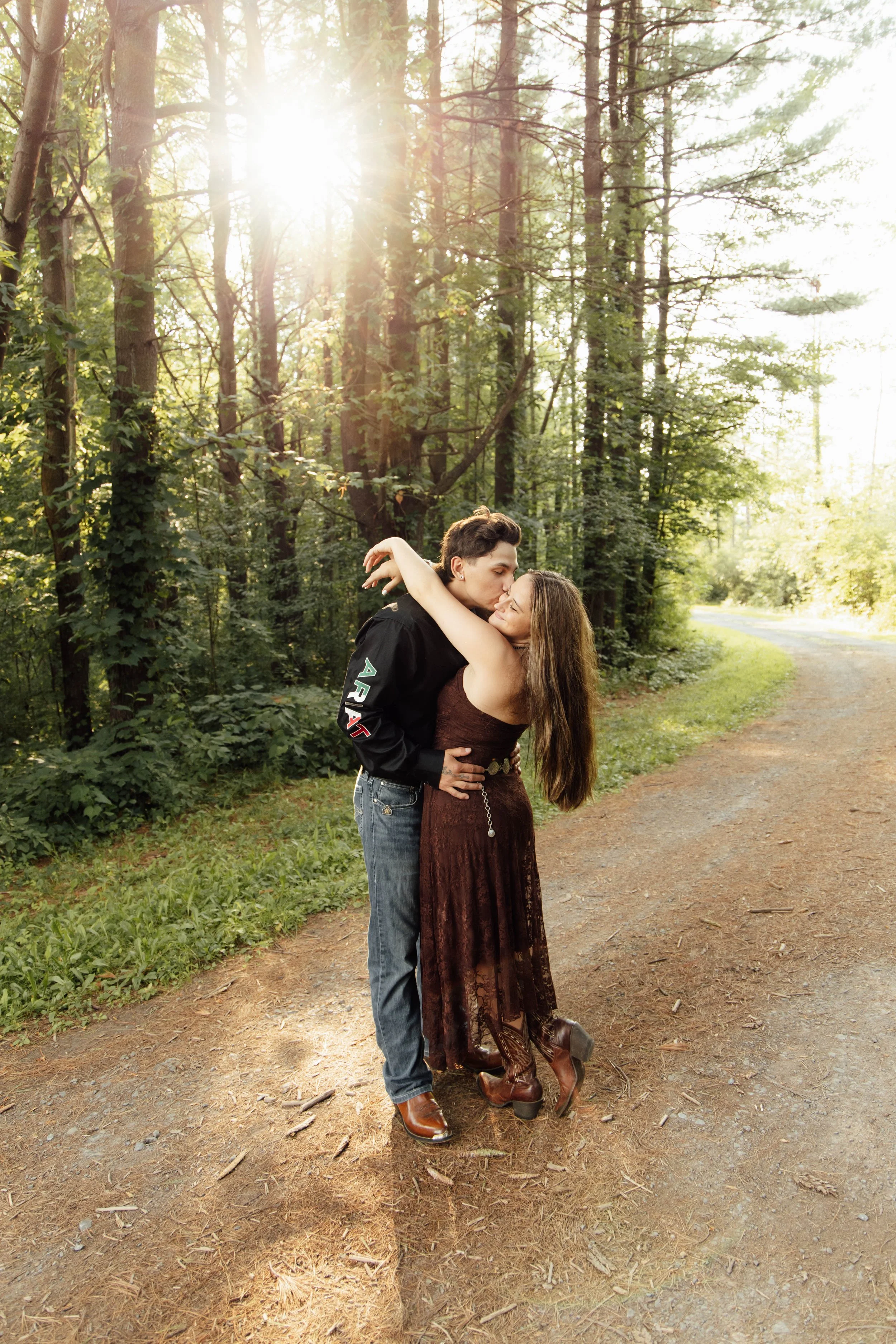A young couple standing close together on a dirt path in a forest, embracing and kissing with sunlight shining through the trees.