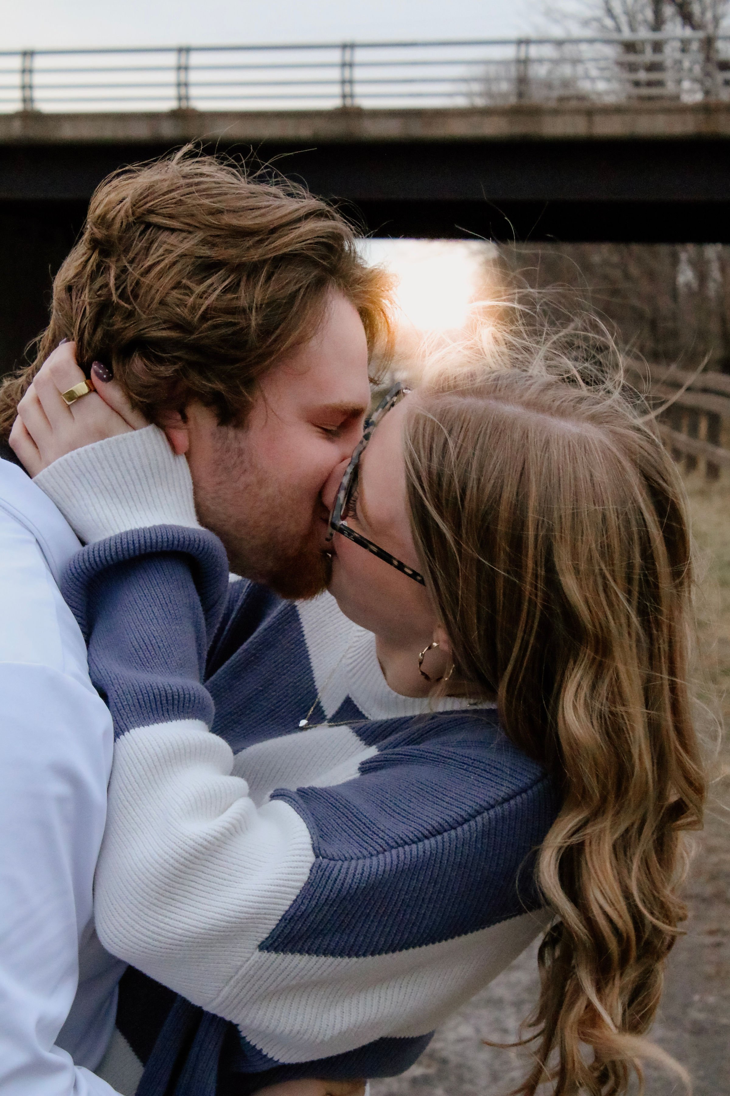 A young couple kissing outdoors under a bridge during sunset.