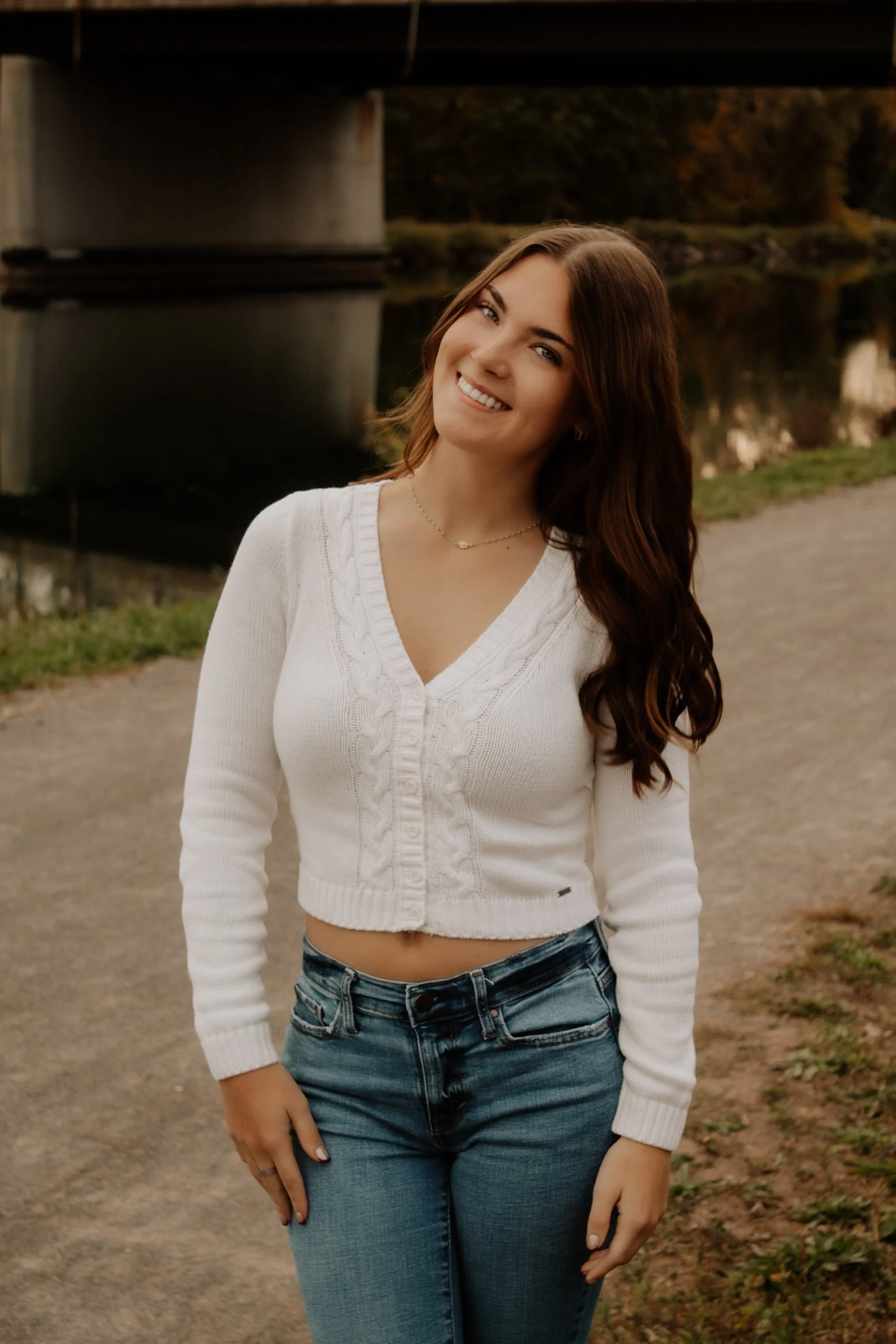 A young woman with long wavy brown hair, wearing a white V-neck cable knit sweater and blue jeans, standing outdoors near a body of water with a bridge overhead, smiling at the camera during sunset.
