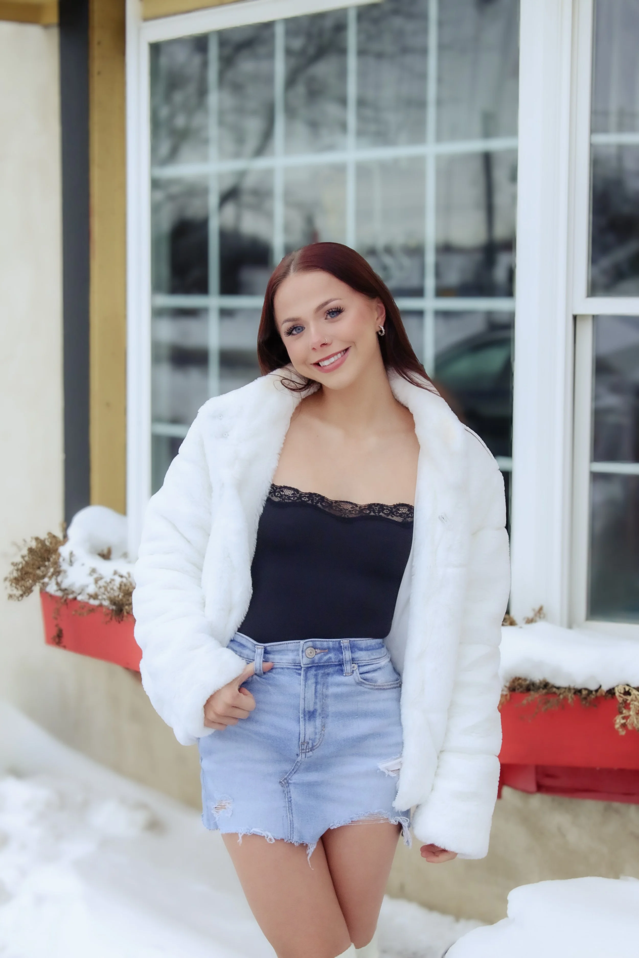 Young woman with red hair smiling outdoors in winter, wearing a white fur coat, black lace top, and ripped denim skirt, standing beside a snow-covered window box.