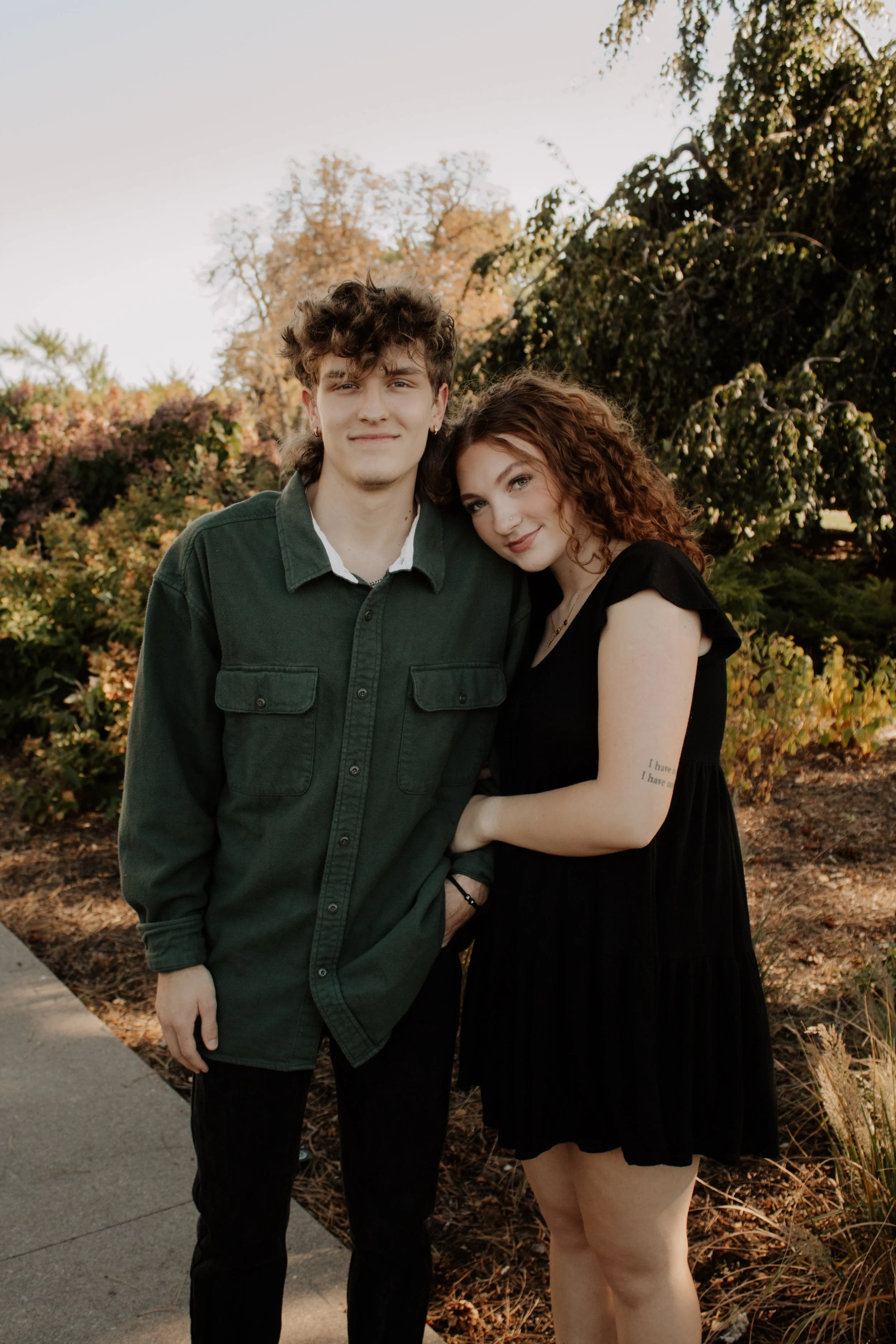 A young man and woman standing close together outdoors in a park during autumn. The man has curly brown hair and is wearing a green jacket over a white shirt. The woman has long, curly red hair and is wearing a black dress with a tattoo on her arm. T