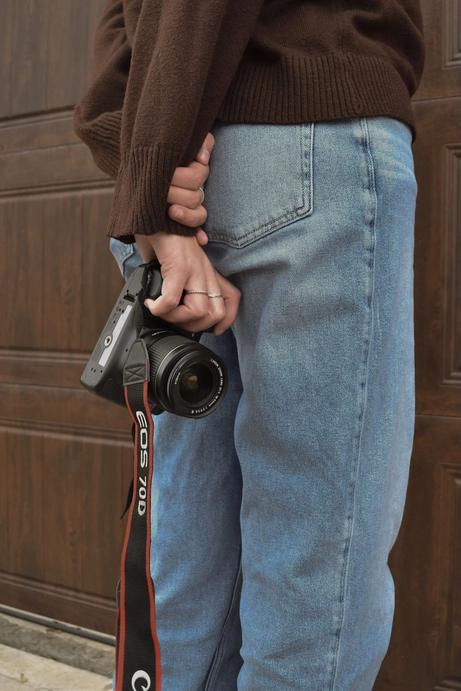 Person holding a Canon camera in their right hand, wearing a brown sweater and light blue jeans, standing in front of a wooden door.