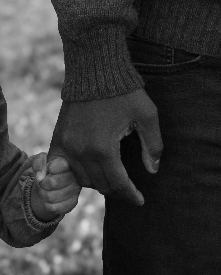 An adult hand holding a child's hand, both in dark clothing, outdoors on a blurred background.