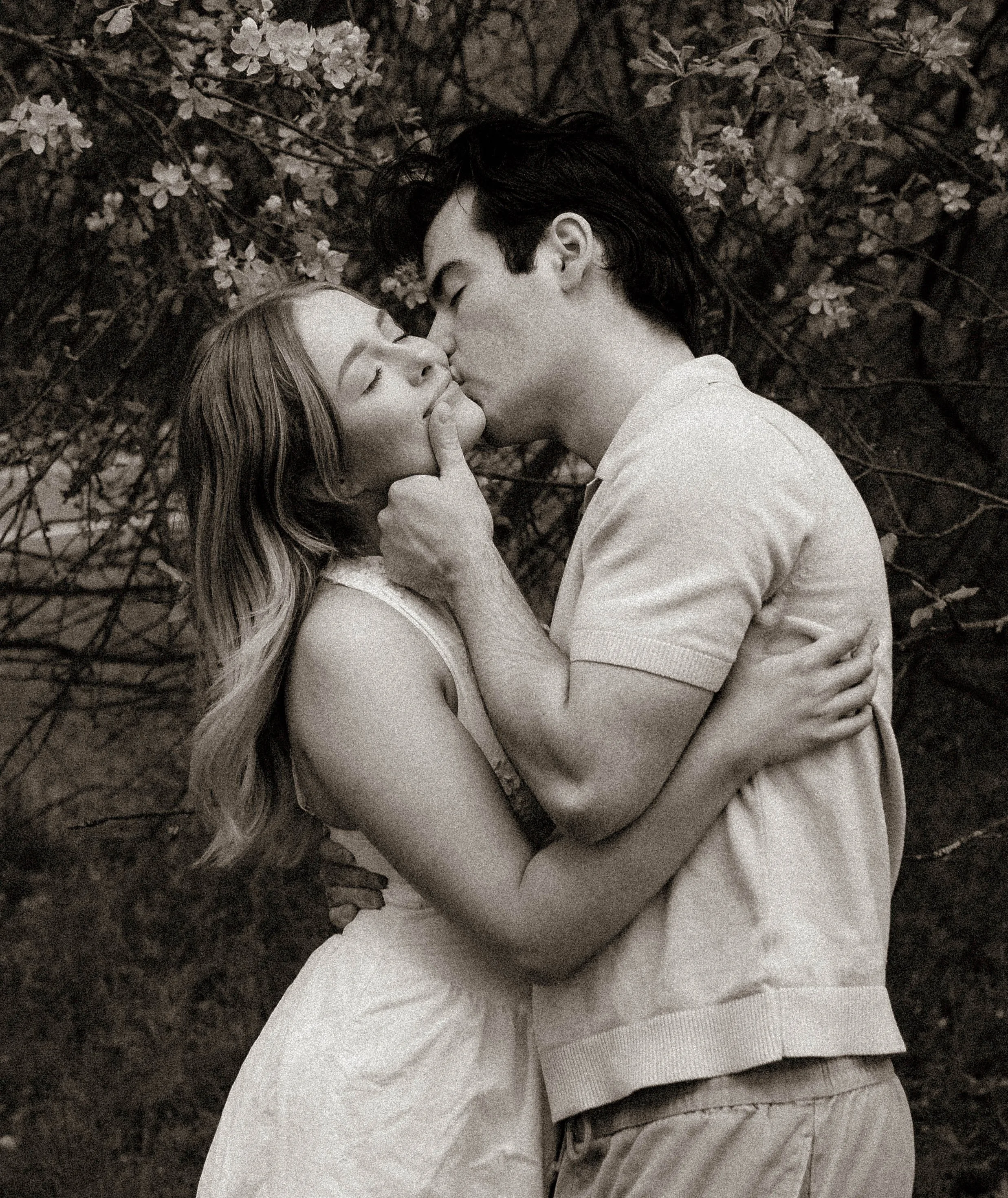 A black and white photo of a young couple kissing, with the man's hand gently touching the woman's face, surrounded by trees and blooming flowers.