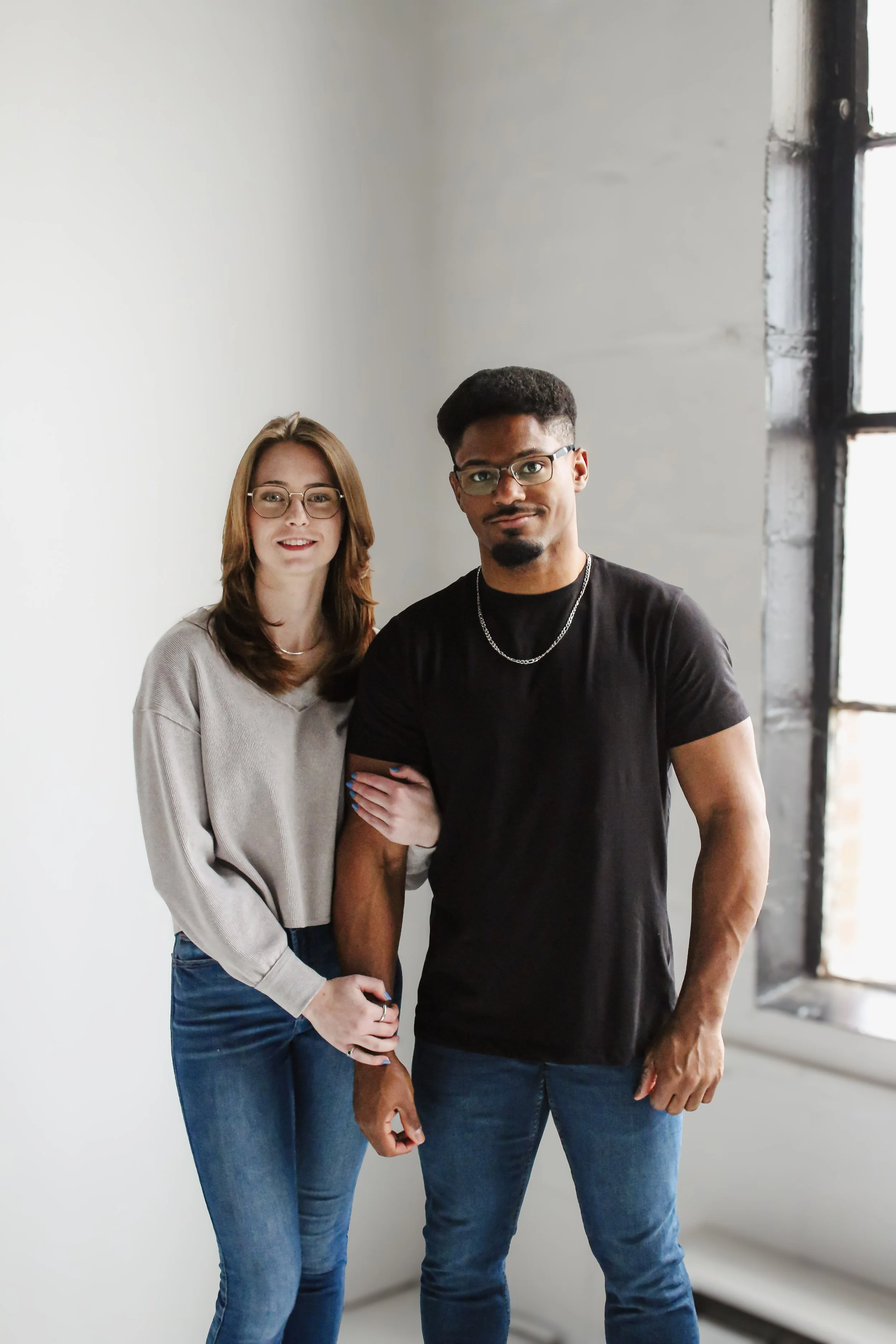 A young woman with brown hair, wearing glasses, a beige sweater, and blue jeans, standing next to a young man with curly hair, glasses, a black t-shirt, and blue jeans, in a brightly lit room with a large window.