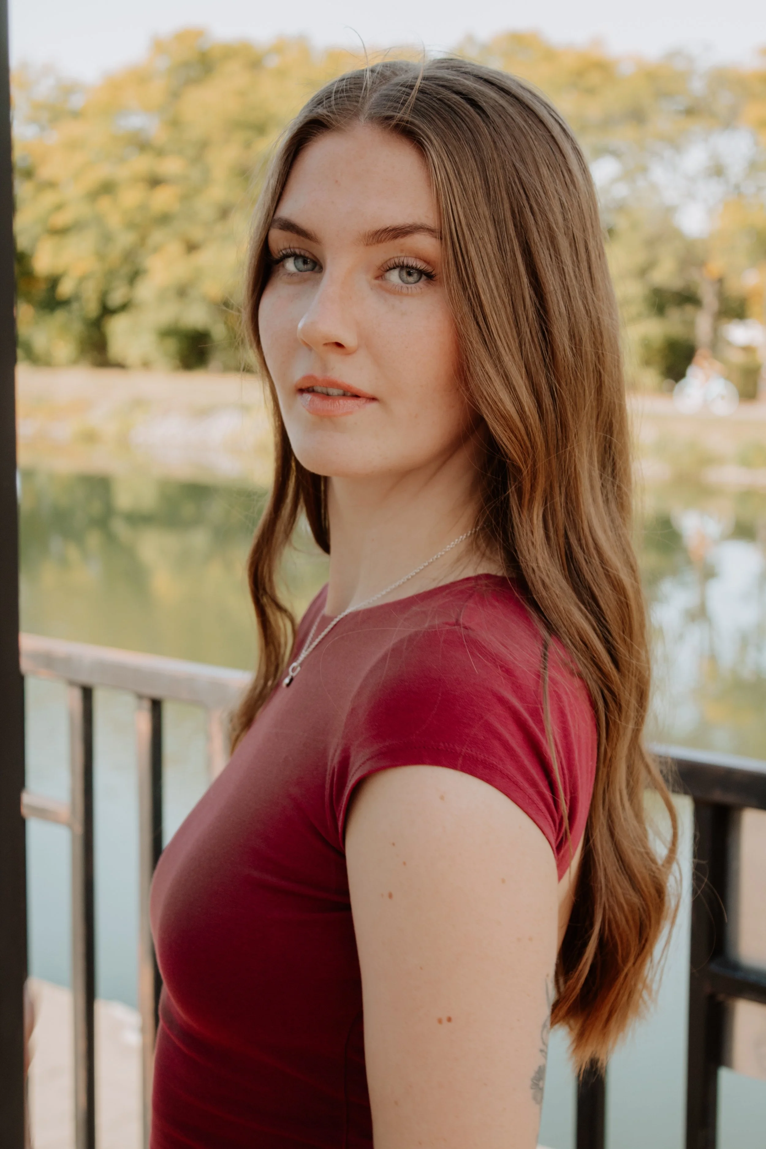 A young woman with long red hair, blue eyes, and light freckles, wearing a red top and a silver necklace, standing outdoors by a railing near a body of water with trees in the background.