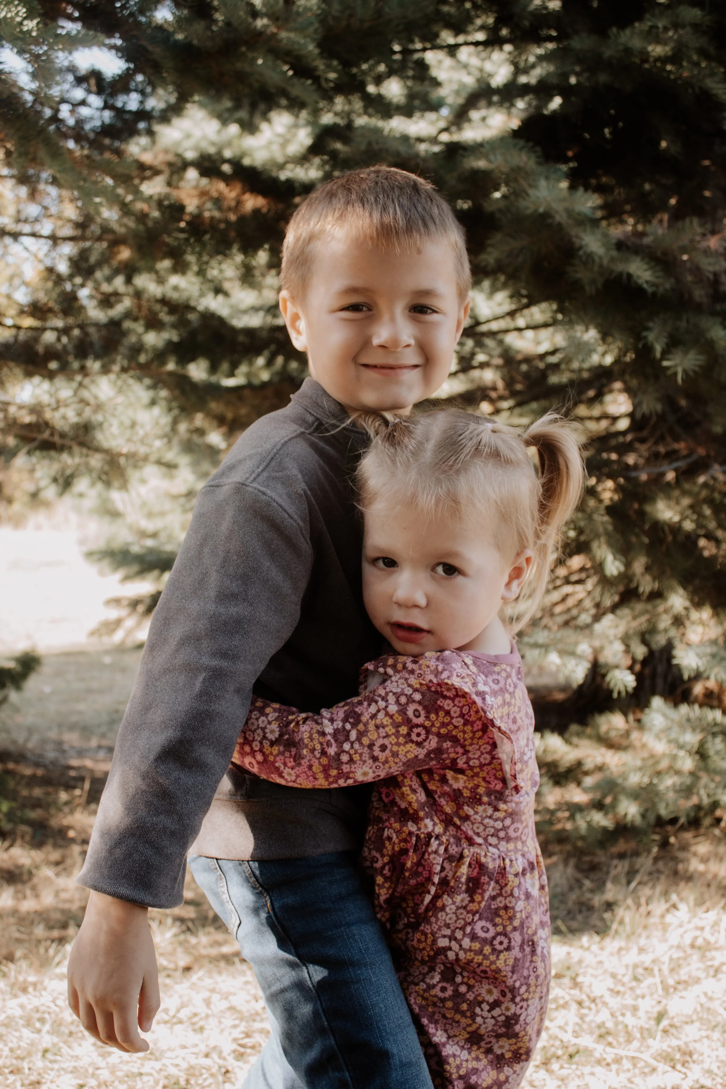 A young boy and girl hugging outdoors near a pine tree, smiling at the camera.