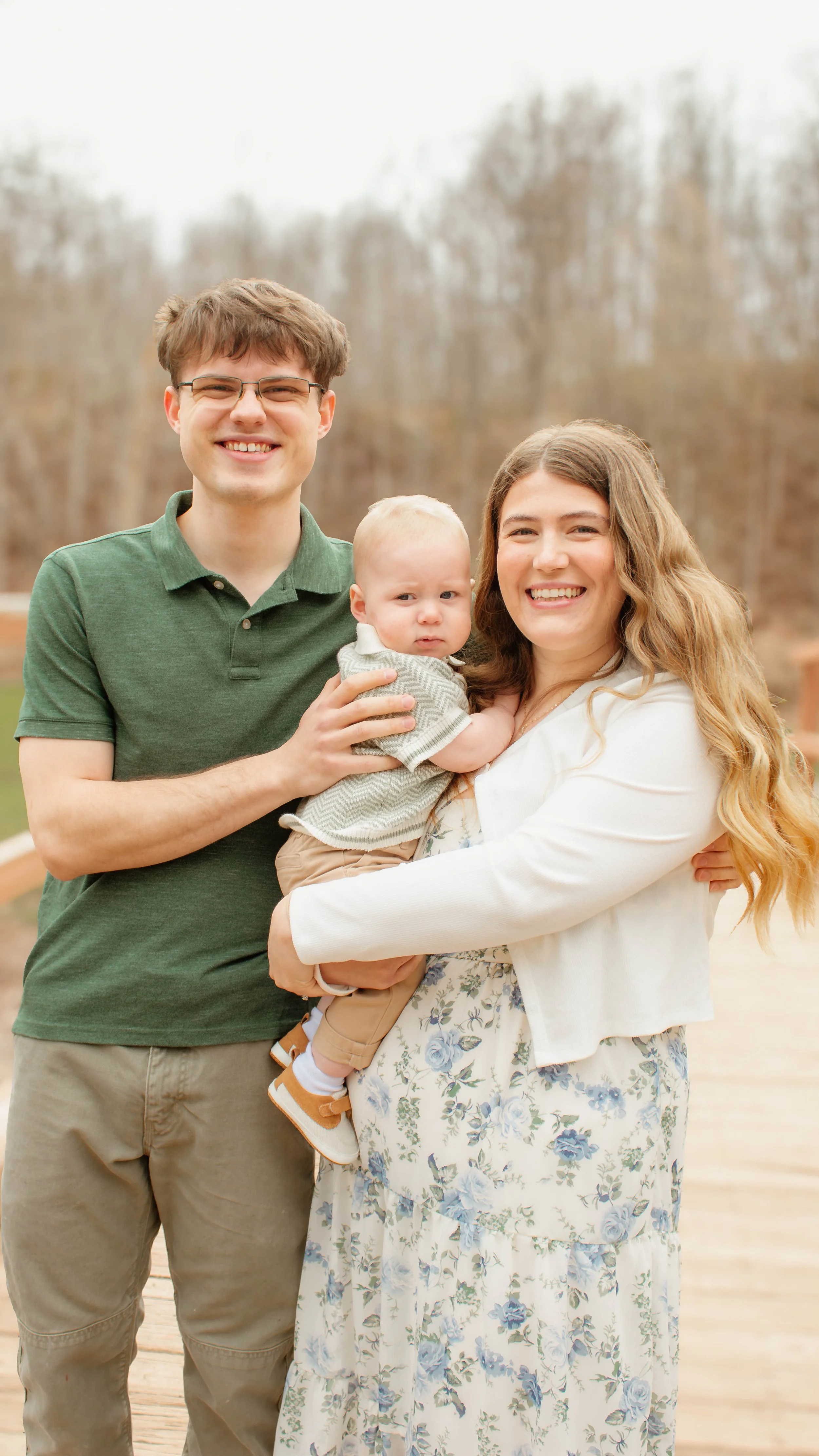 A smiling family of three with a man, woman, and baby, standing outdoors on a cloudy day with trees in the background, on a wooden walkway.