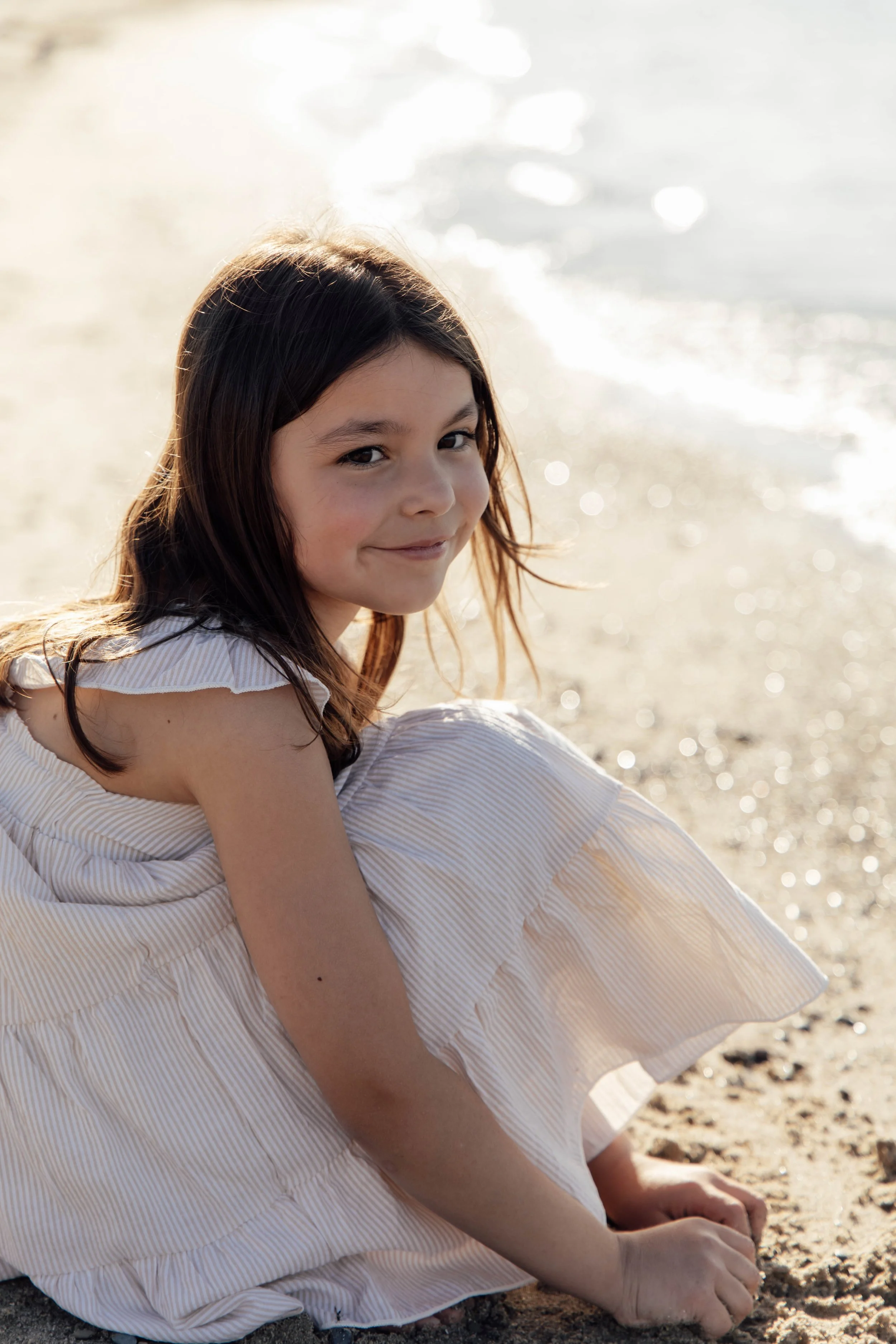 A young girl with long, dark hair sitting on the beach, smiling slightly, wearing a white and beige striped dress, with the sun reflecting off the water in the background.