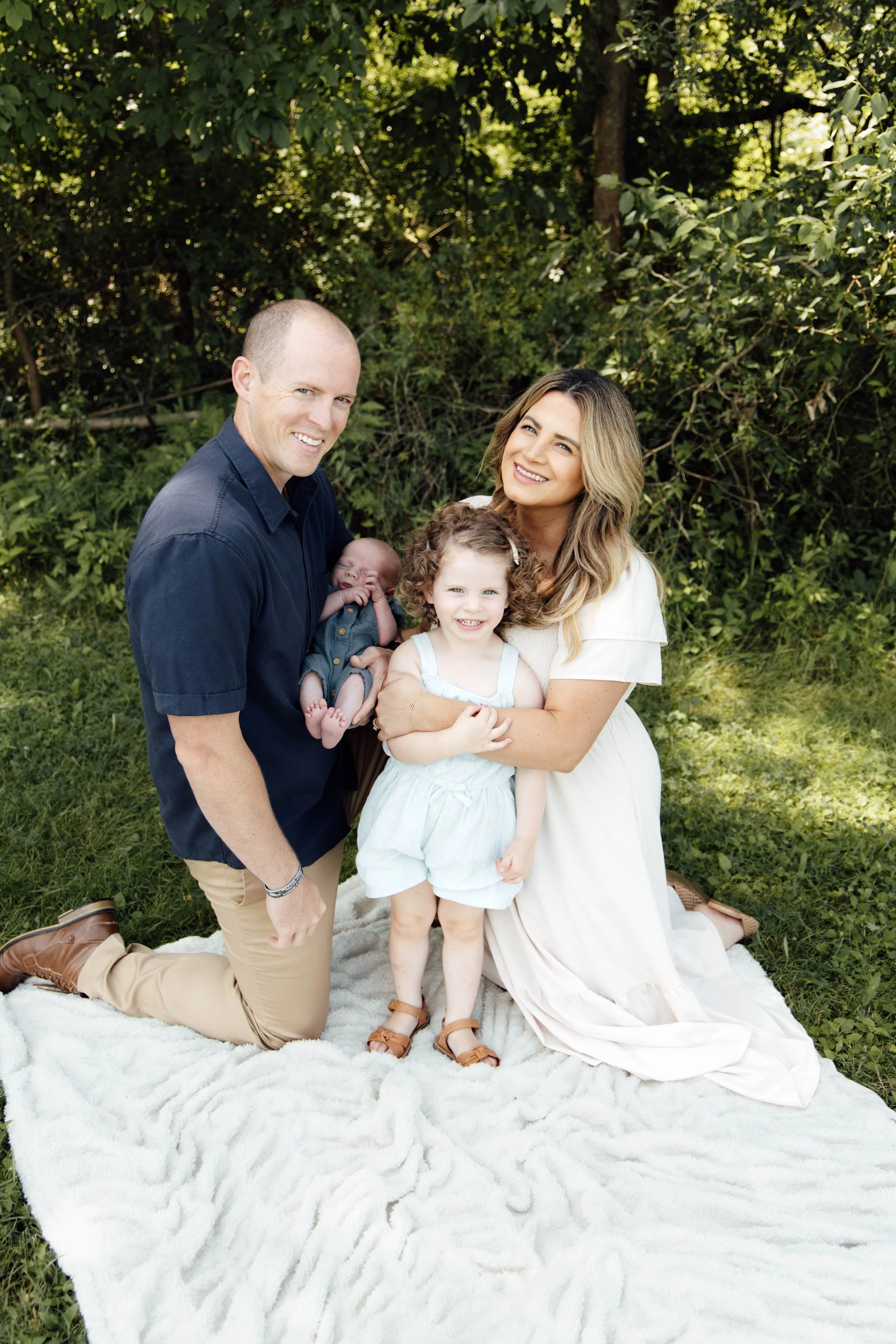 A family of four posing outdoors on a grass lawn with green trees in the background. The father, kneeling on a white blanket, is smiling and holding a newborn baby. The mother, sitting on the blanket, is smiling and hugging a young girl with curly hair. All are dressed casually and appear happy.