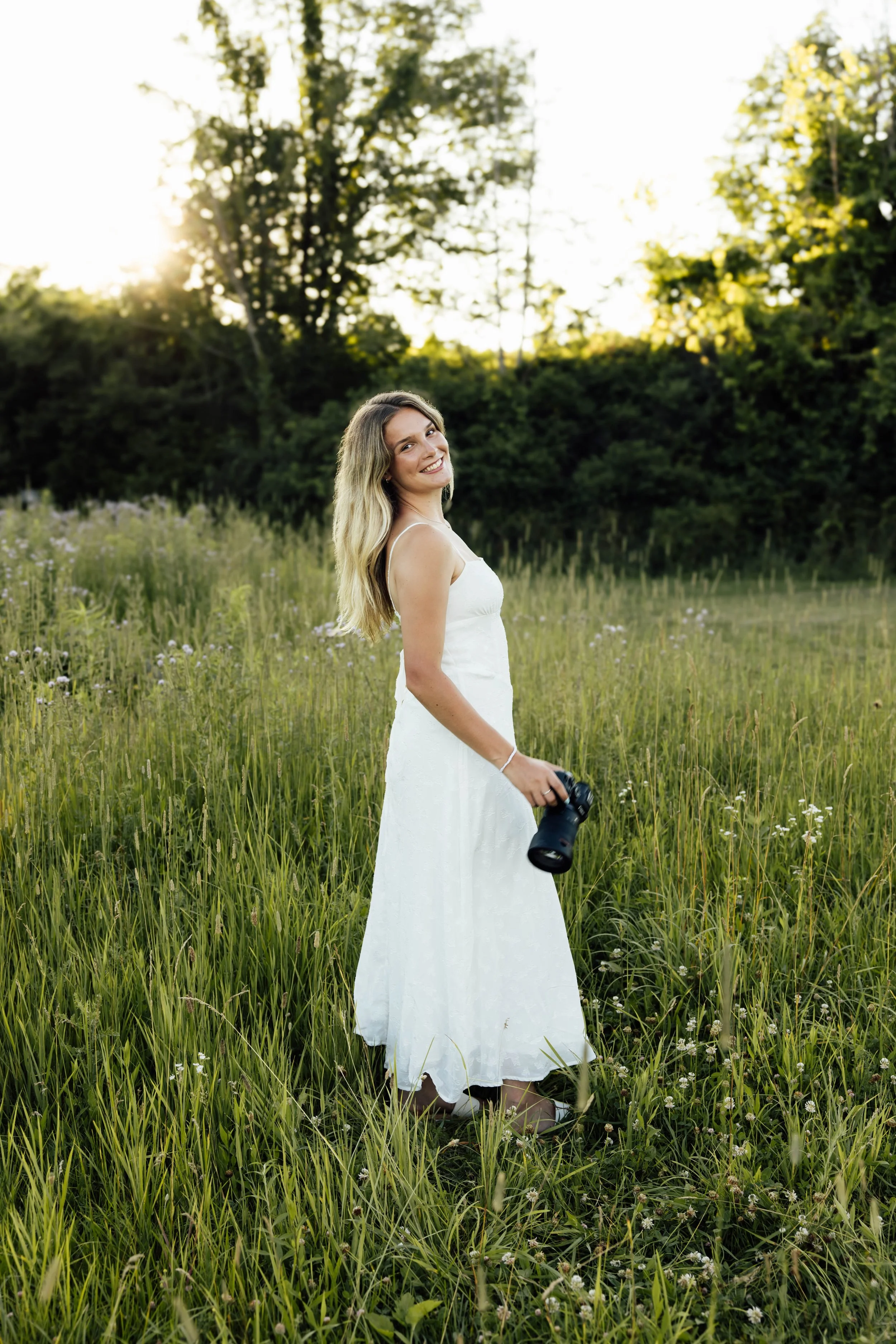 A young woman in a white dress standing in a grassy field, holding a camera and smiling at the camera, with trees and setting sunlight in the background.