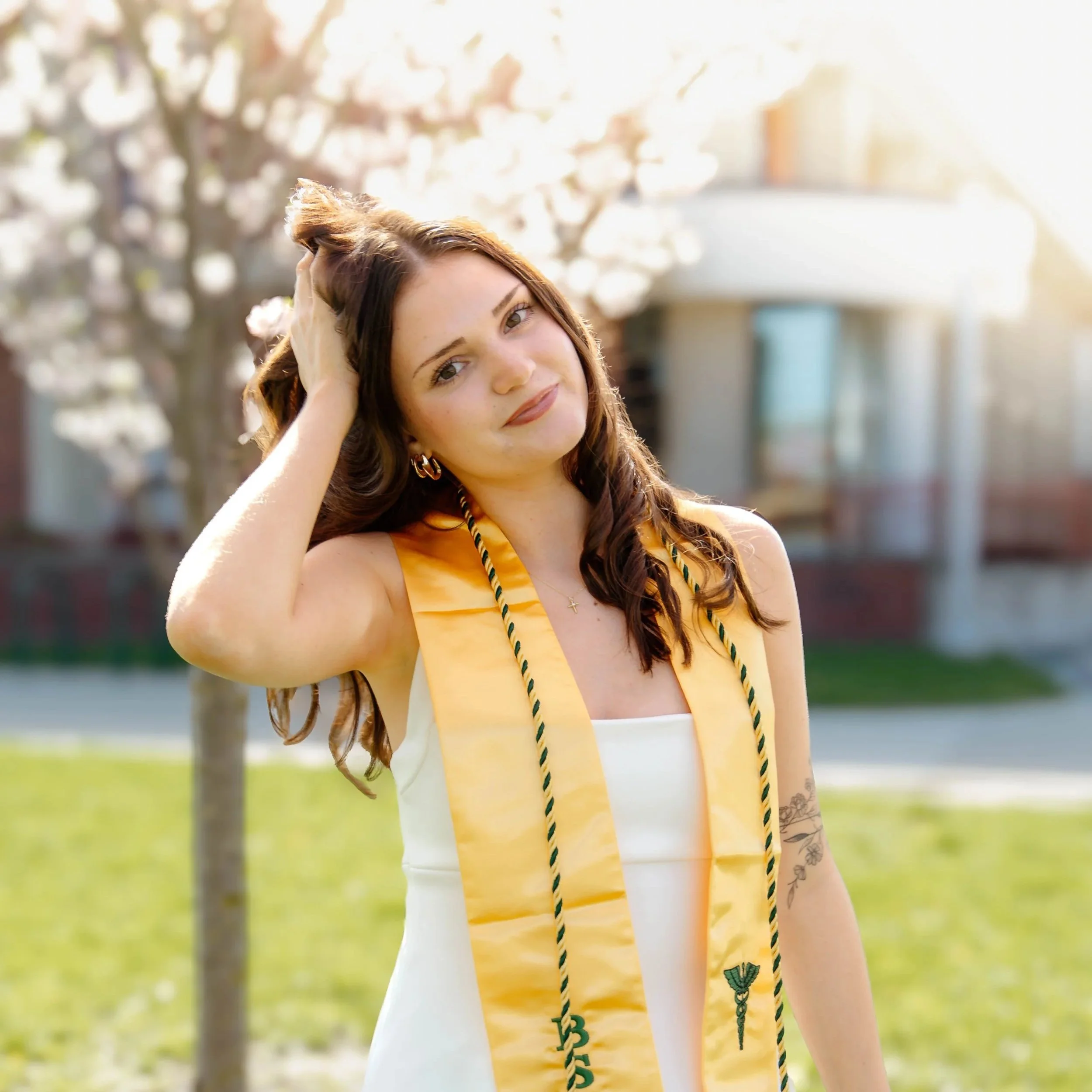 A young woman with long, wavy brown hair, wearing a yellow graduation stole over a white dress, standing outdoors in daytime with sunlight. She has a small tattoo on her left arm and is holding her hair with her right hand, smiling softly.