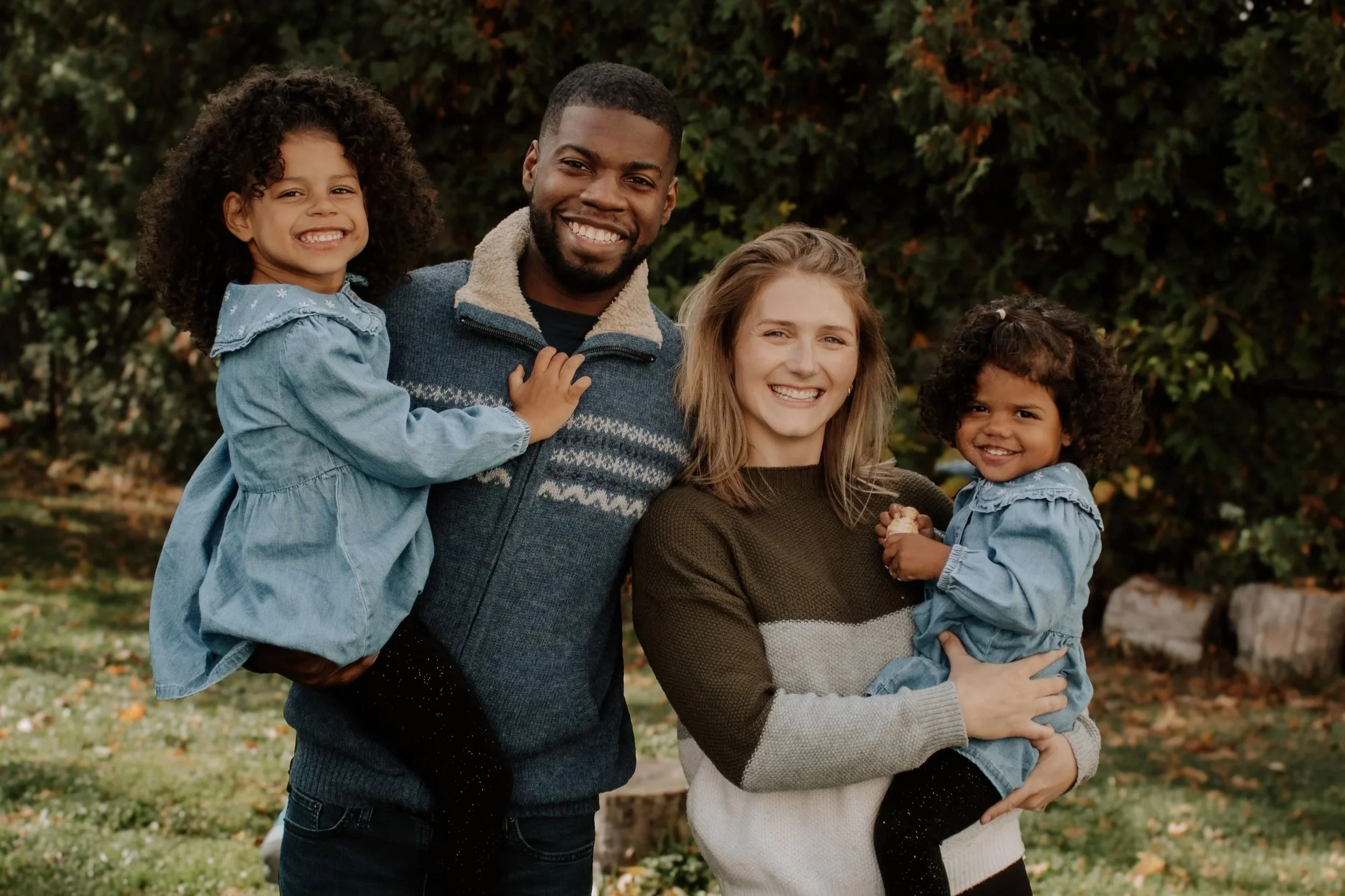 A diverse family with two women and two children smiling outdoors in a park or garden.