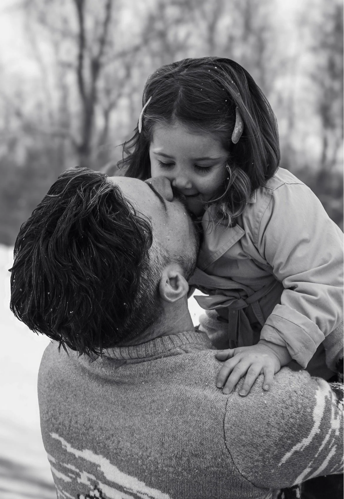 A man and a girl share a kiss outdoors; the man is holding the girl, who is leaning in to kiss him, with a background of blurred trees.