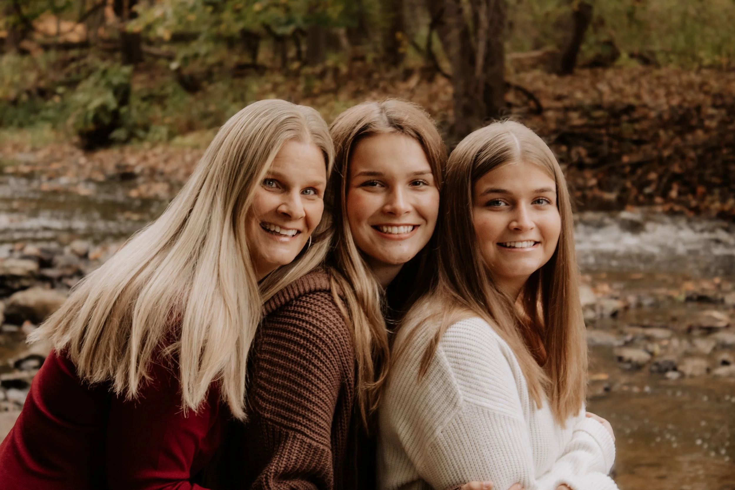 Three women with long hair smiling outdoors near a river with rocks and trees in the background.