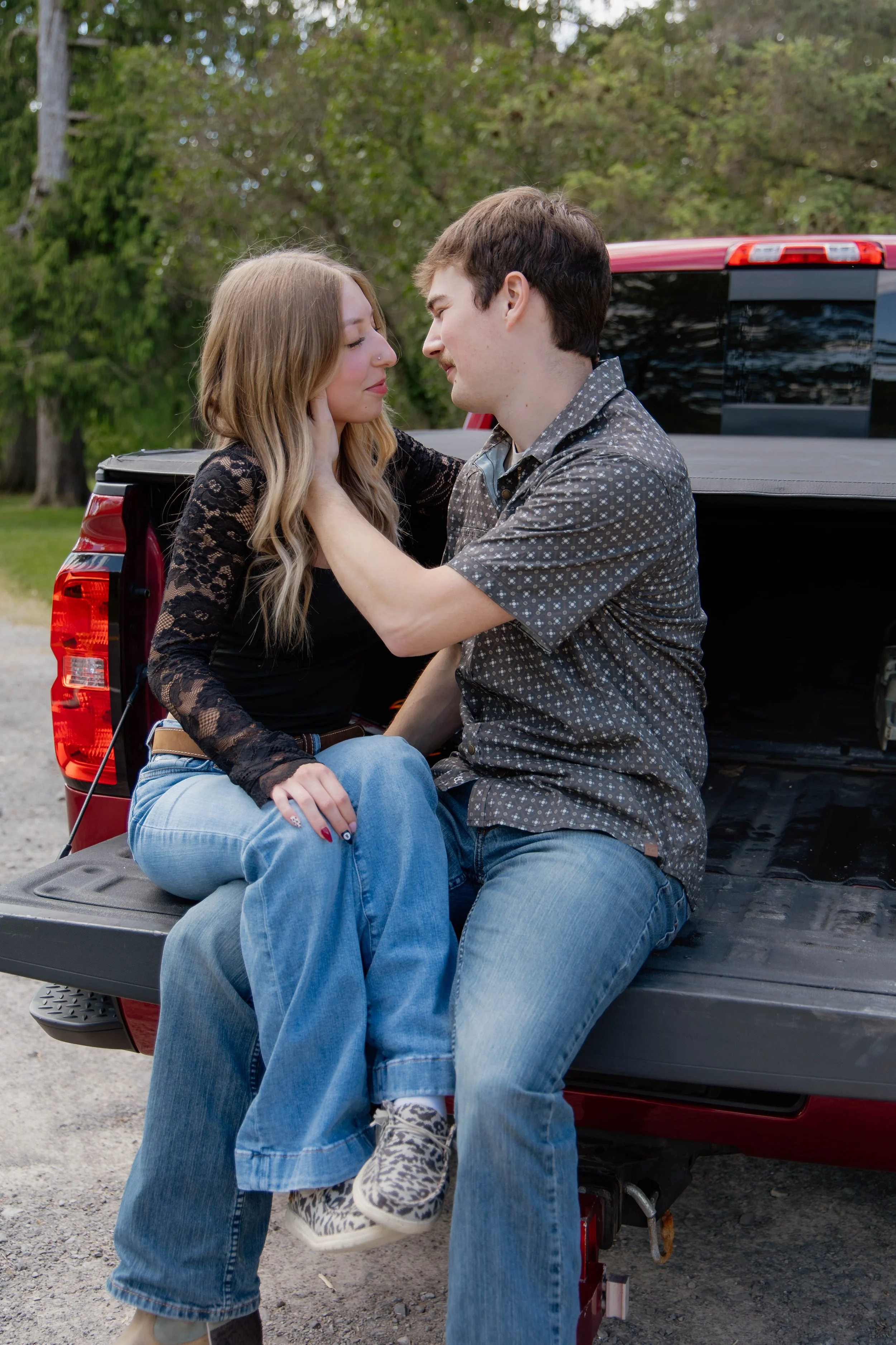 A young couple sitting on the tailgate of a red pickup truck outdoors, with trees and greenery in the background. The woman is sitting on the man's lap, and they are looking into each other's eyes, smiling gently.