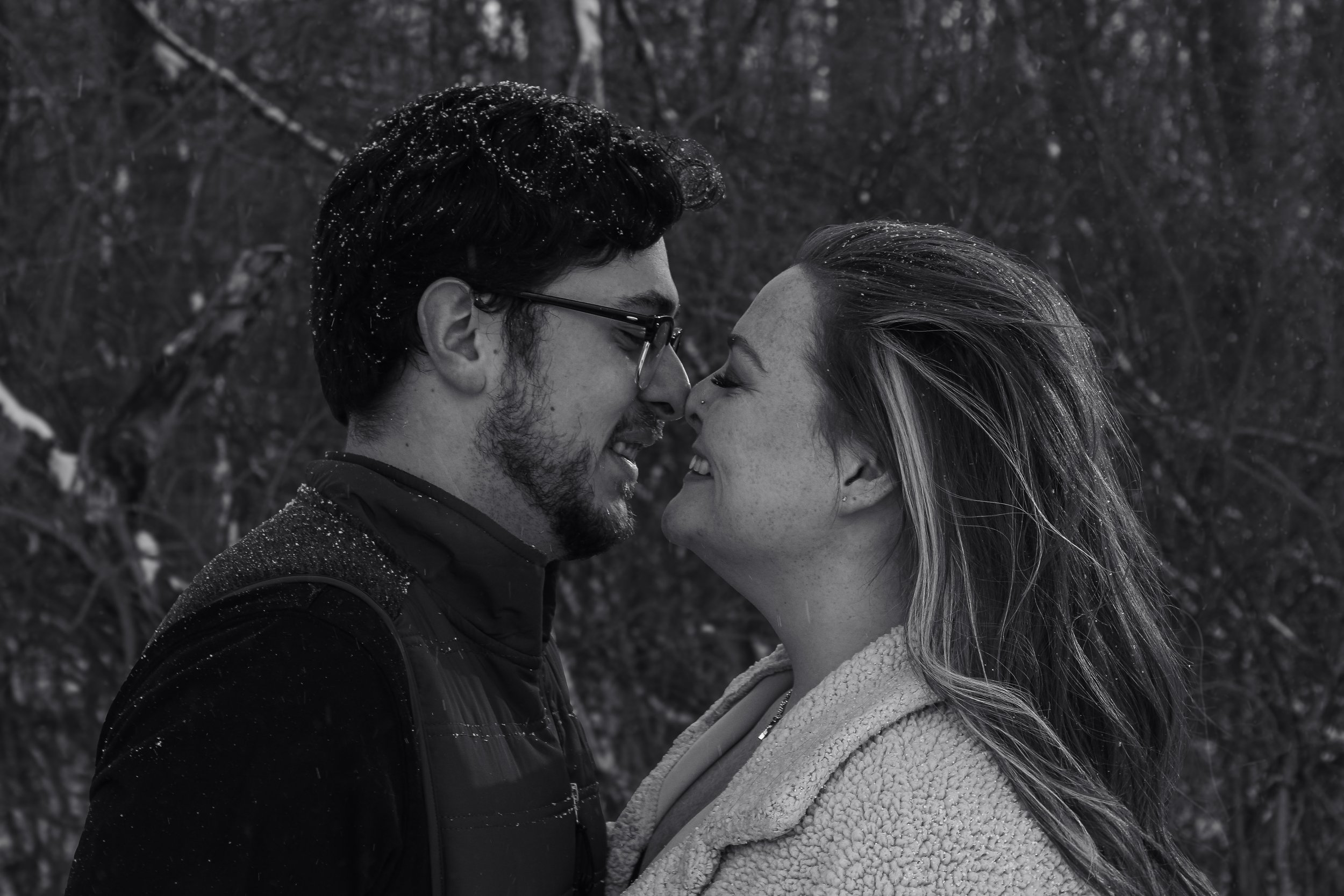 A black and white photo of a man and woman standing close, touching foreheads and smiling in a snowy outdoor setting.