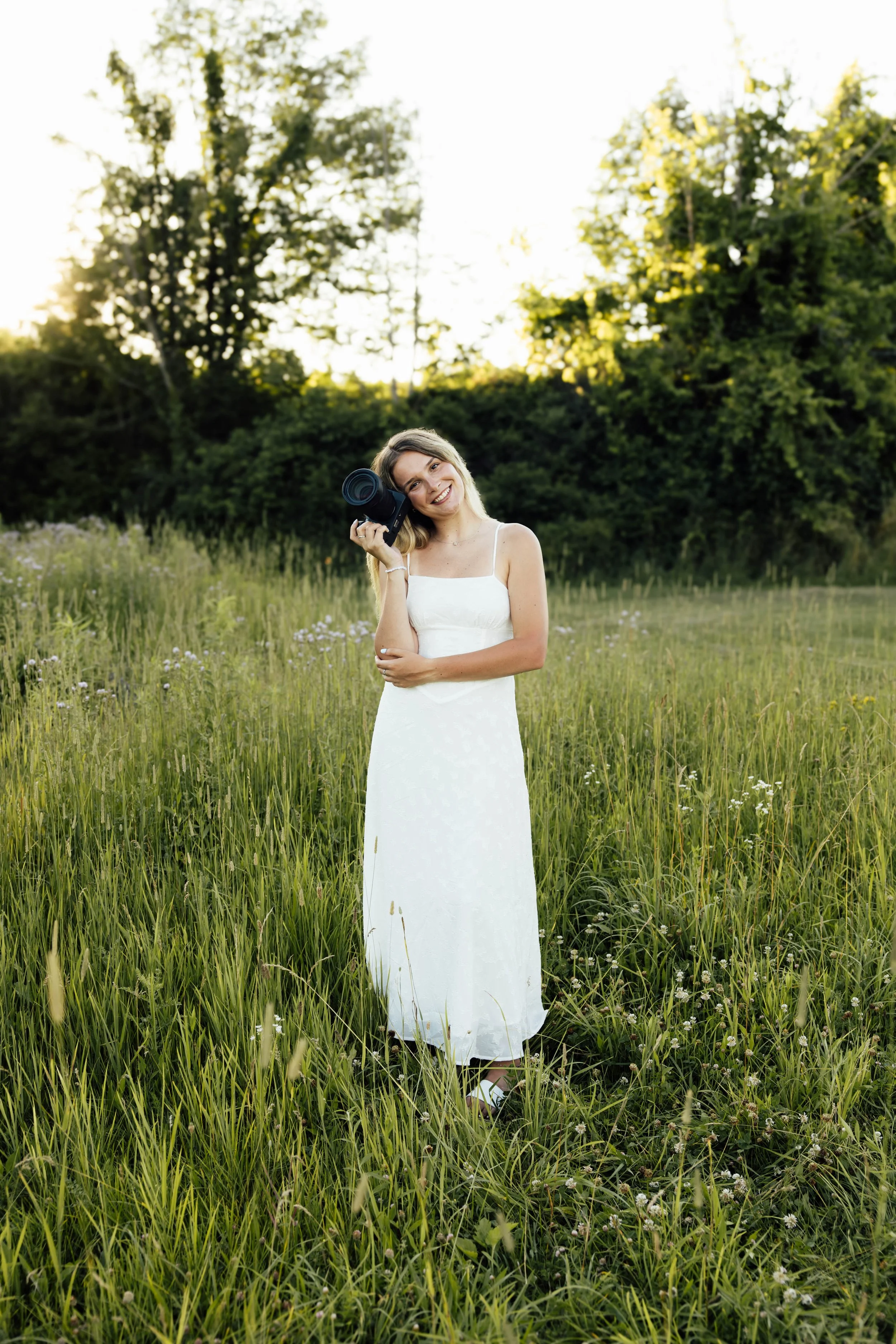 A woman in a white dress standing in a grassy field holding a camera. She is smiling with her head tilted and is surrounded by tall grass, wildflowers, trees, and the sun setting behind the trees.