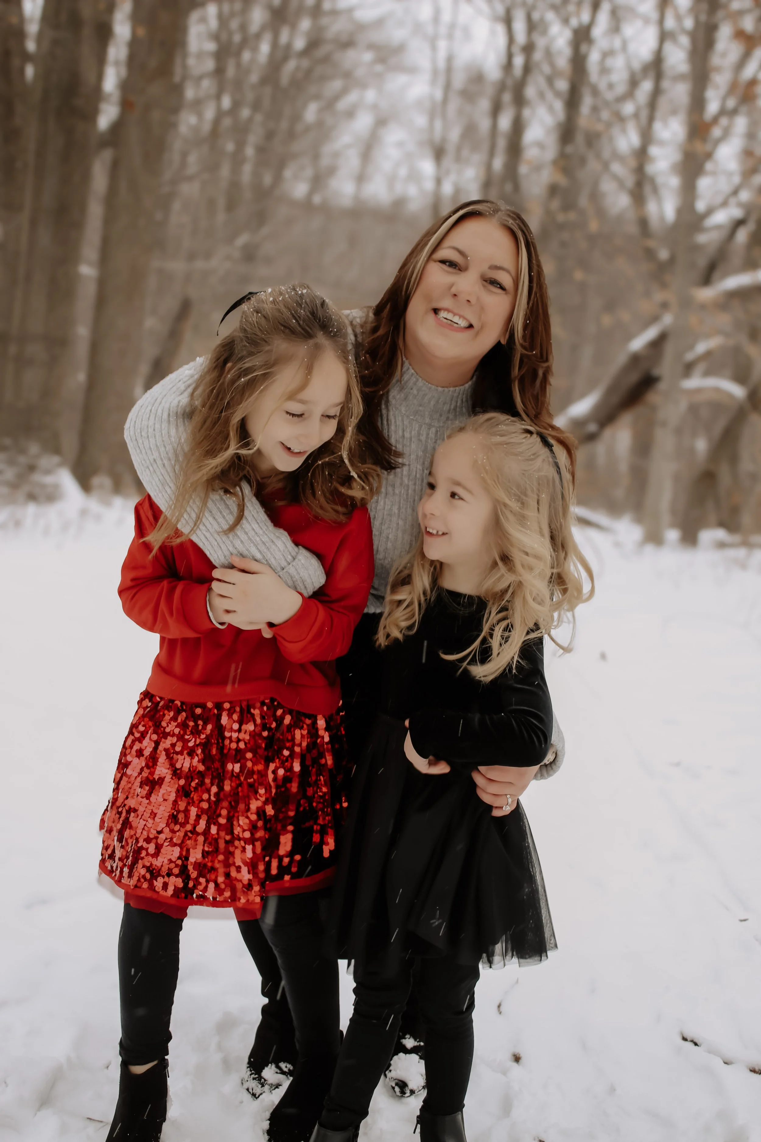 A woman with two young girls standing outdoors in a snowy wooded area, all smiling and hugging each other.