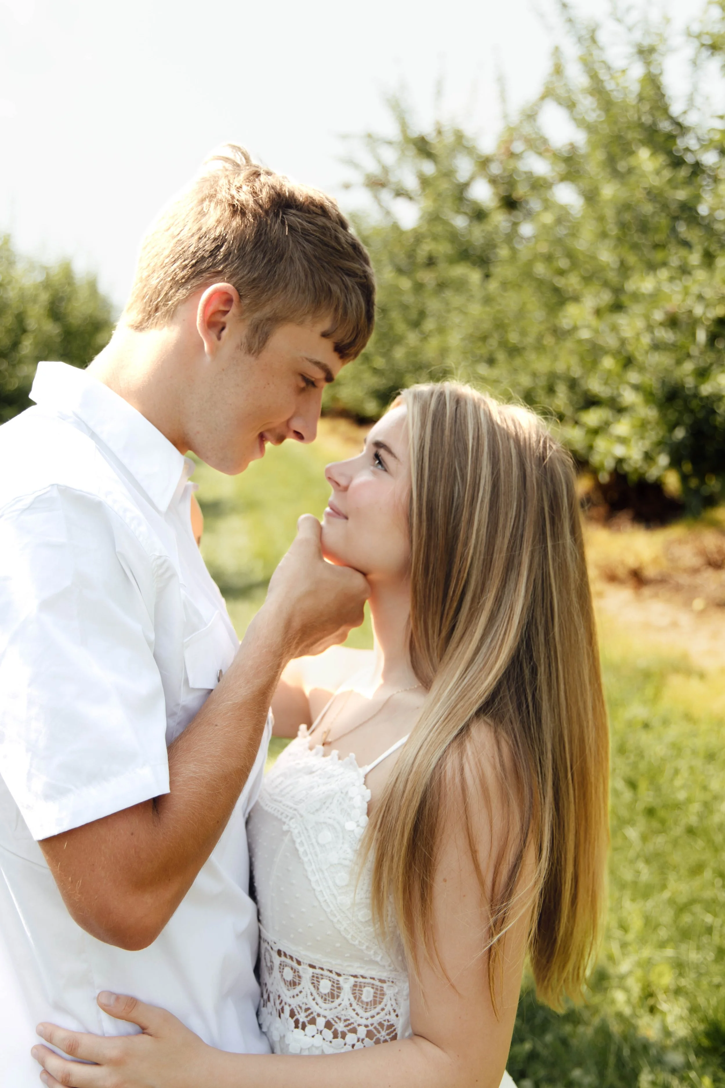 A young couple is outdoors, looking into each other's eyes with affection. The man is gently holding the woman's face with one hand, and they are smiling. The background features green trees and sunlight, creating a warm and romantic atmosphere.