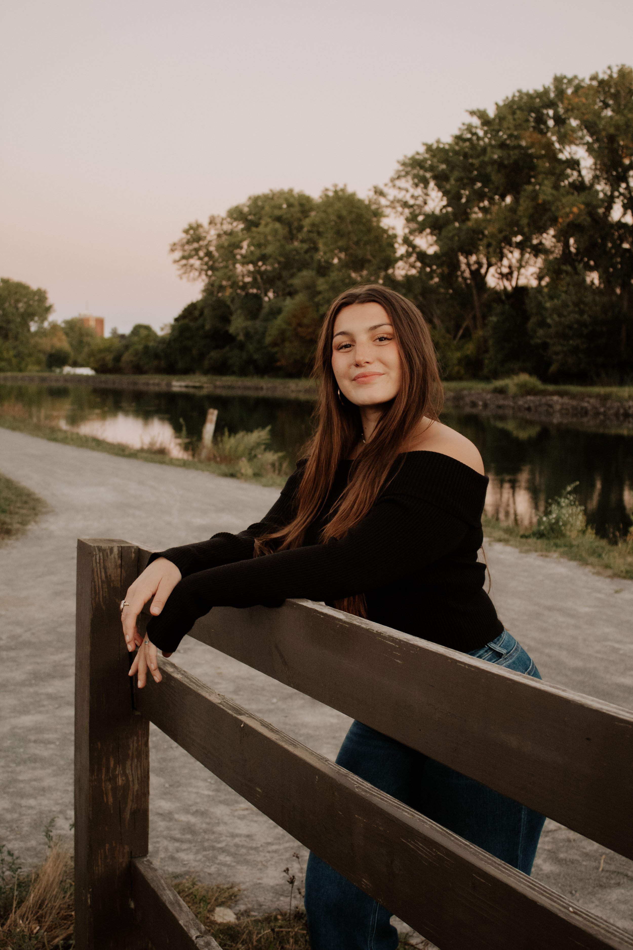 A young woman with long brown hair leaning on a wooden railing outdoors near a river during sunset, wearing a black off-the-shoulder sweater and jeans, smiling softly at the camera.