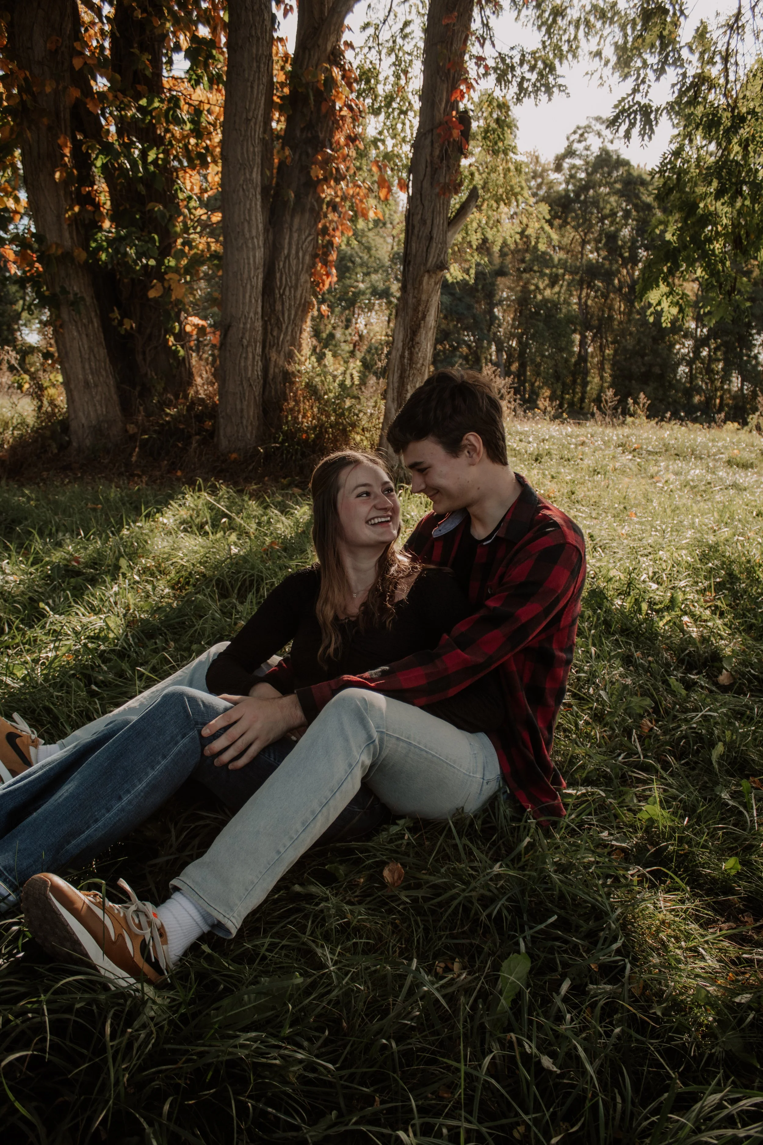 A young couple sitting on the grass in a park, smiling and looking at each other, surrounded by trees with fall foliage.