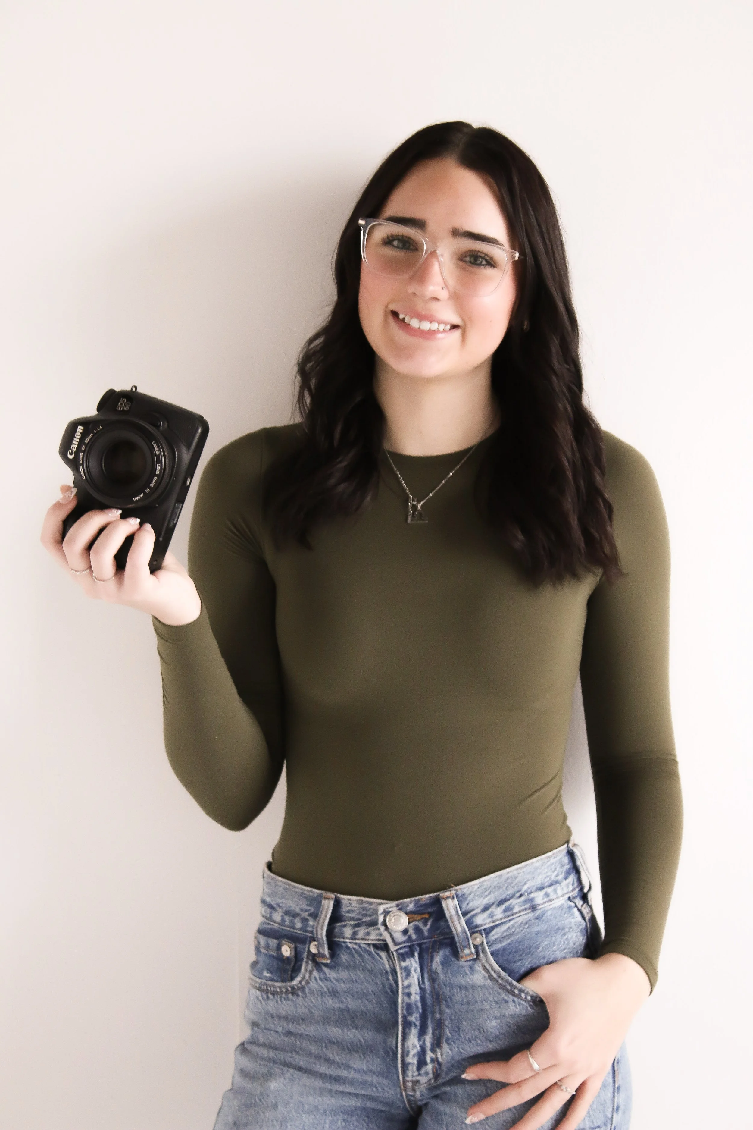A young woman with dark hair, glasses, wearing an olive green long-sleeve top and blue jeans, holding a Canon camera, smiling, standing against a plain white wall.