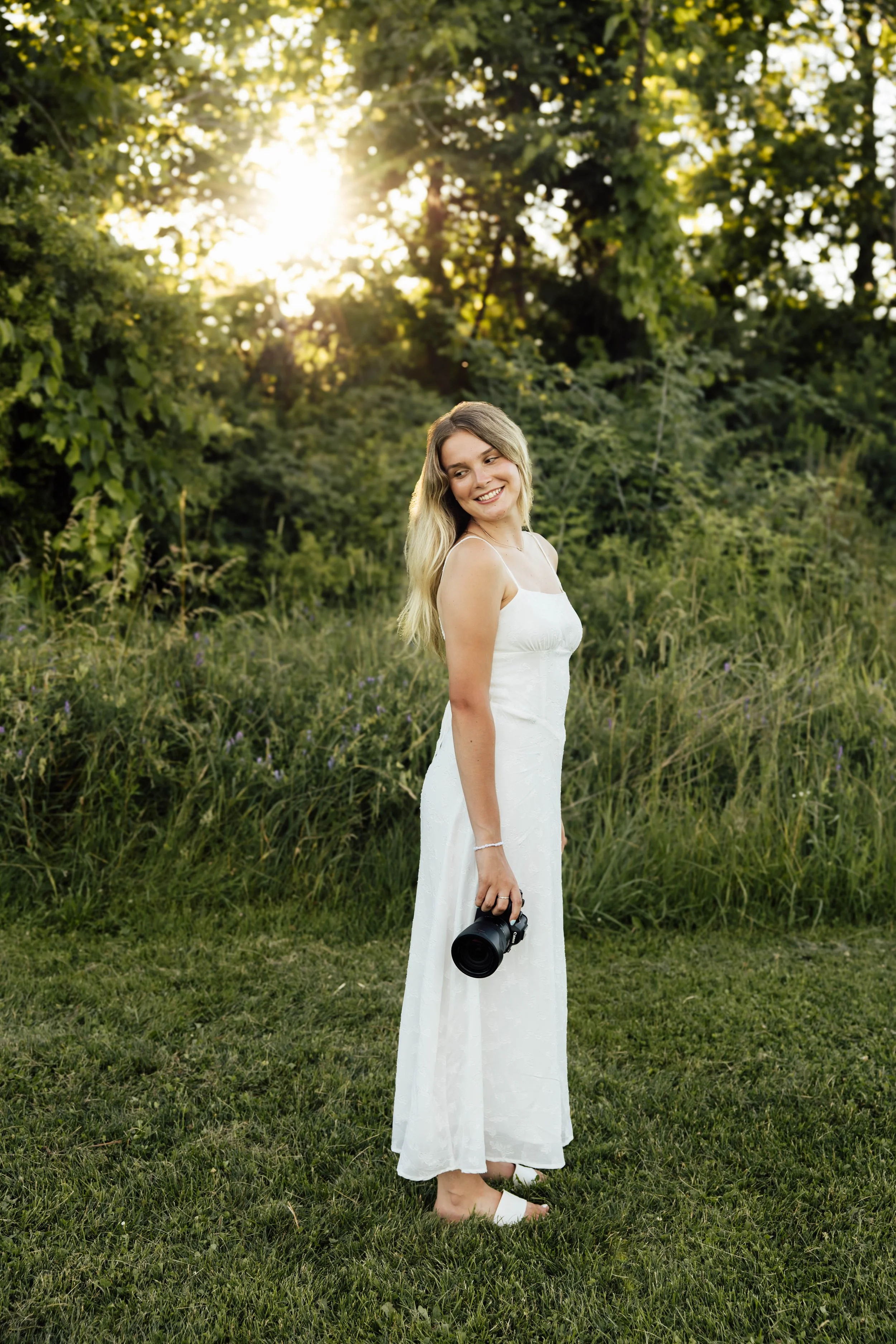 A young woman in a white dress standing outdoors on grass, holding a camera in her right hand, smiling, with trees and sunlight in the background.