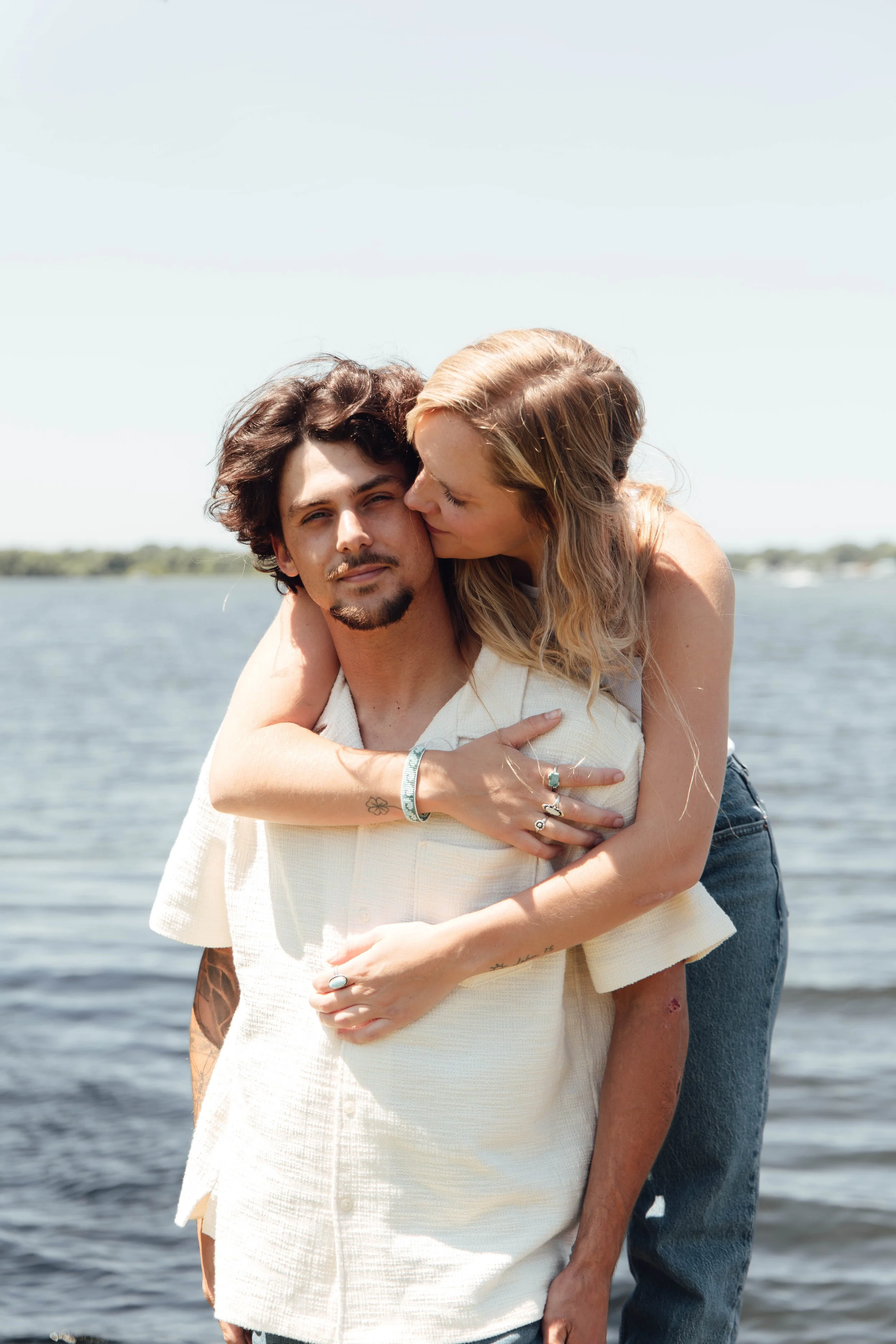 A couple embracing near a body of water with trees in the background on a sunny day.