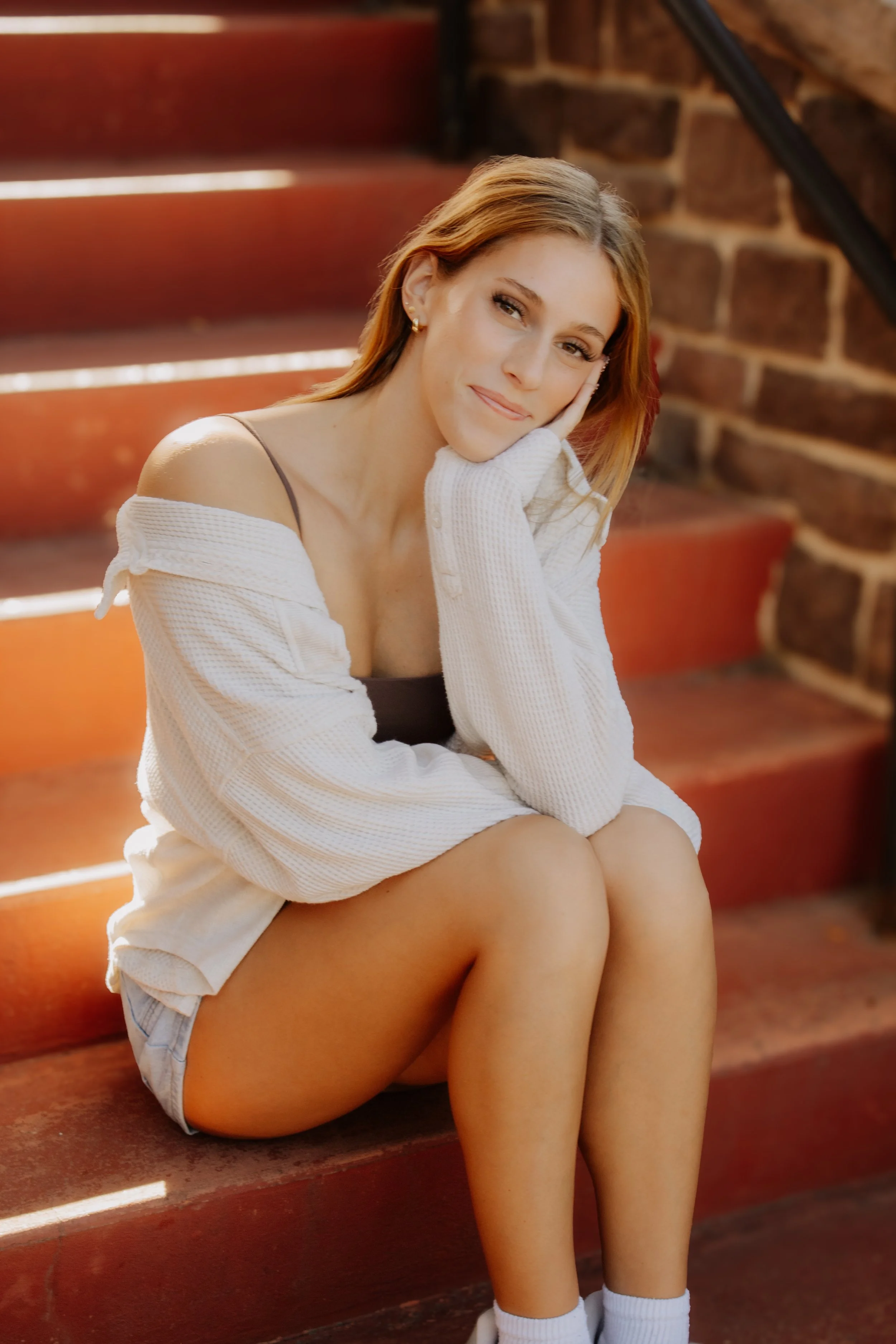 Young woman with shoulder-length hair sitting on red wooden stairs, resting her face on her hand, wearing a white cardigan, black tank top, denim shorts, and white socks.