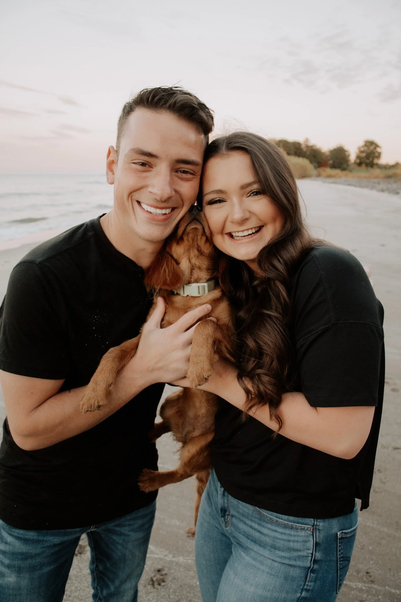 A young couple smiling and holding a small brown dachshund puppy on a beach at sunset.
