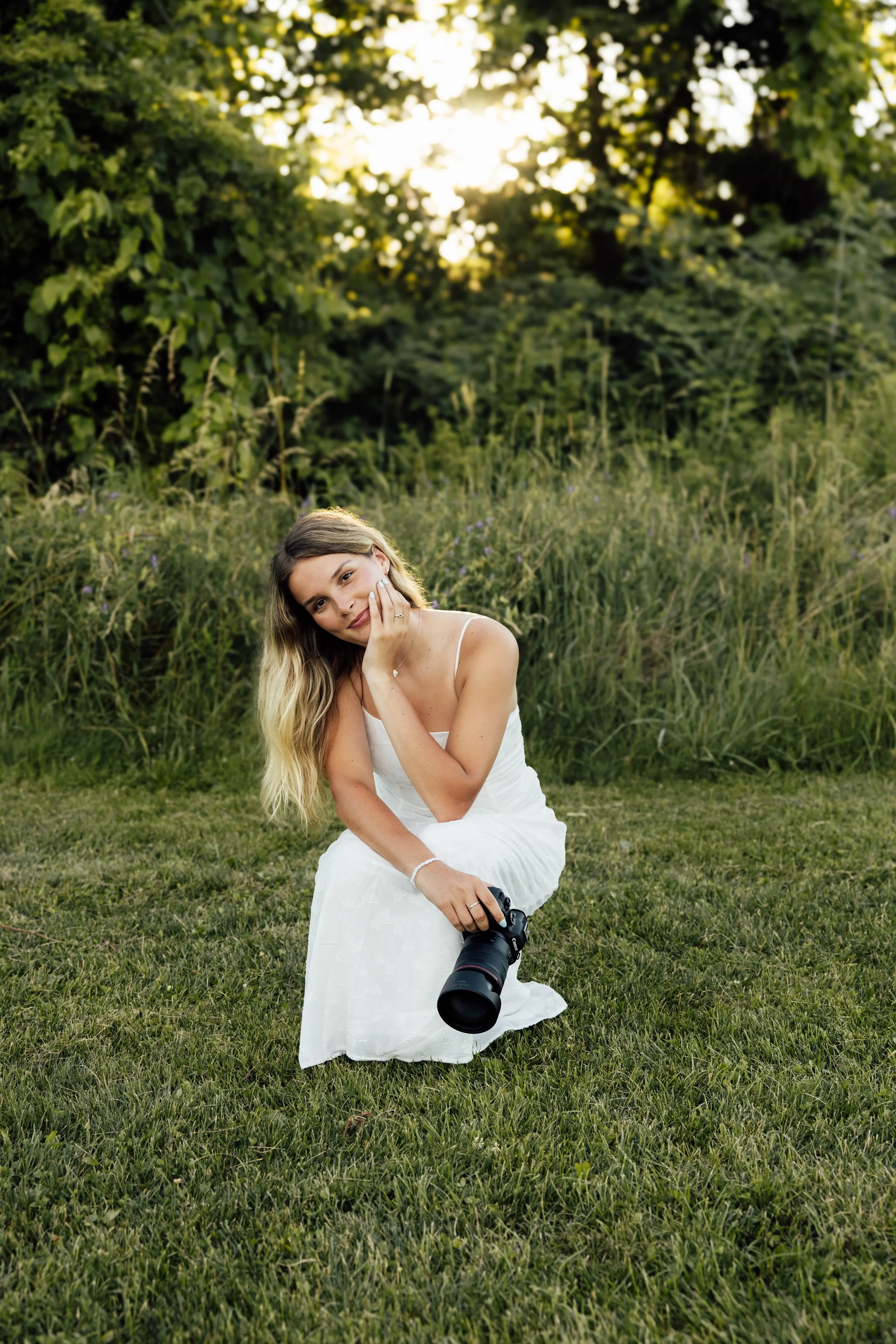 A woman with long blonde hair, dressed in a white dress, crouching on the grass in a field with tall grass and purple flowers. She is holding a professional DSLR camera with a large lens, and resting her head on her left hand, smiling softly, with trees and sunlight in the background.