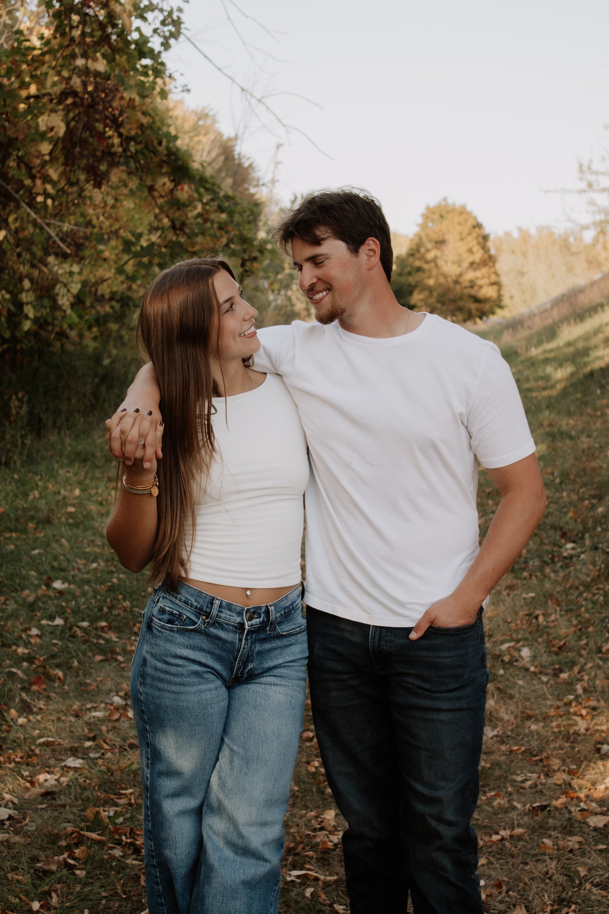 A young couple standing outdoors in a park or nature setting, smiling and looking at each other, with trees and a pathway in the background.
