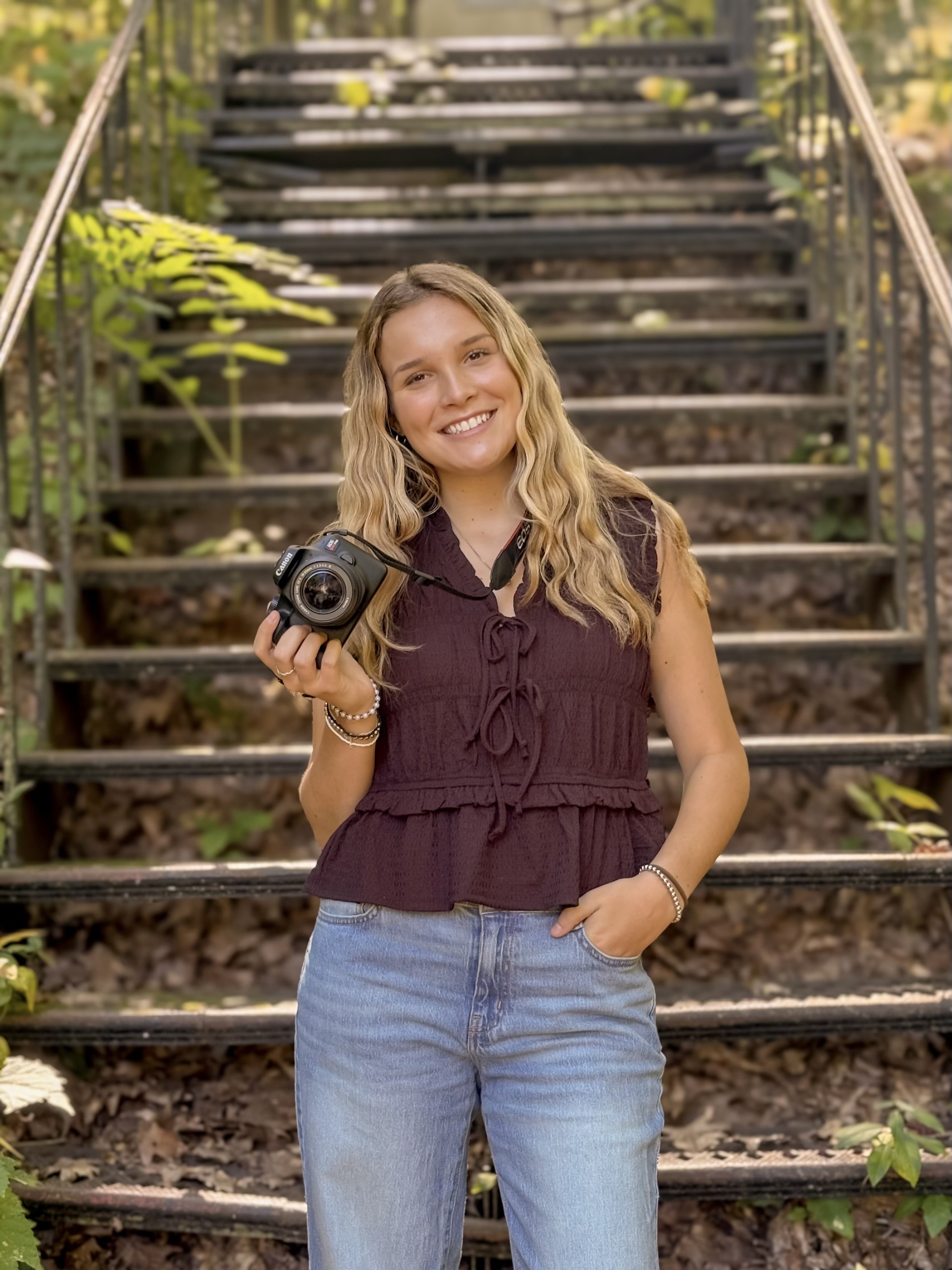 A young woman with long, wavy blonde hair smiling while holding a camera in her right hand, standing outdoors in front of a staircase with greenery around.