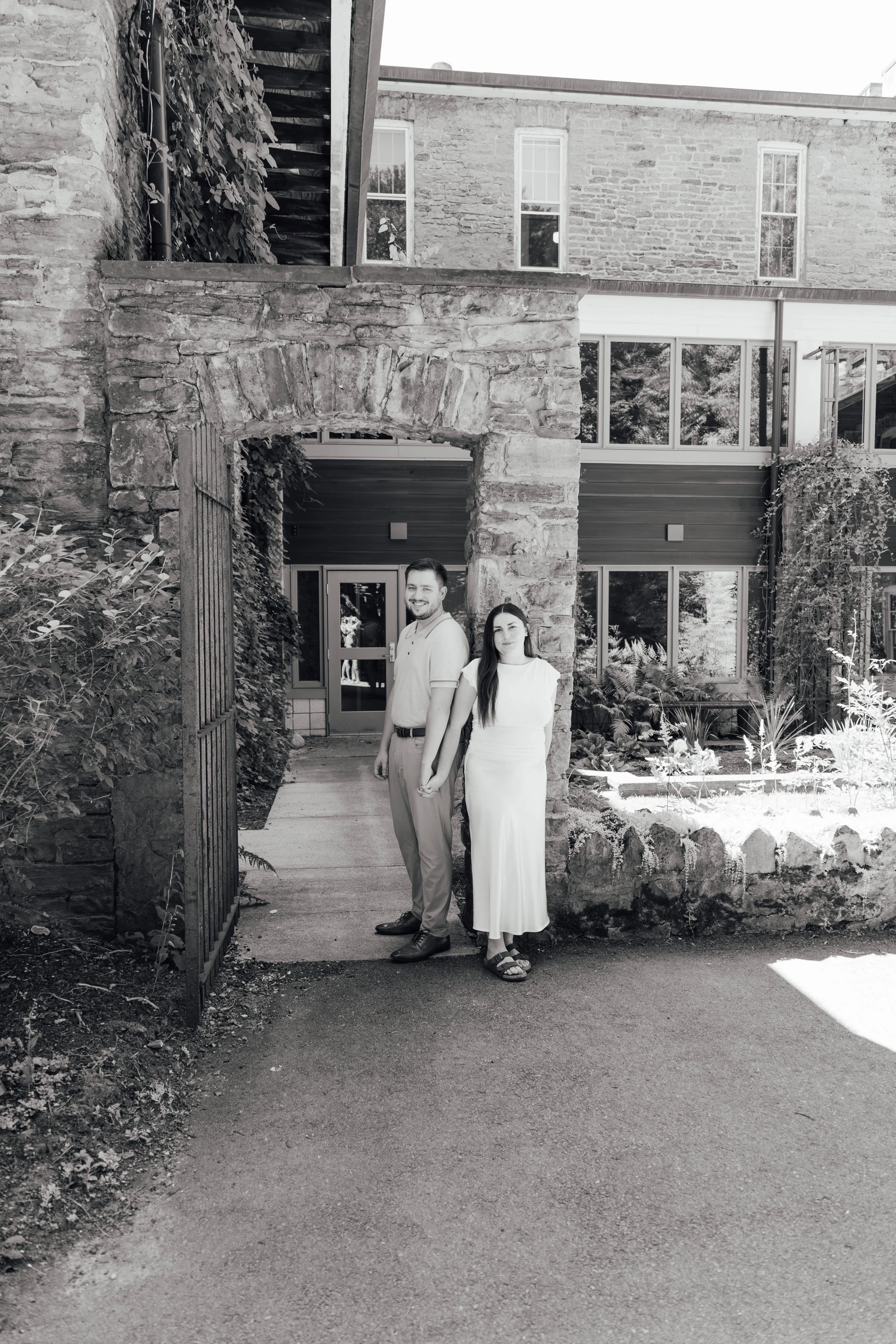 A grayscale photo of a man and woman standing outdoors under a stone archway, holding hands and smiling, with a brick building and greenery in the background.