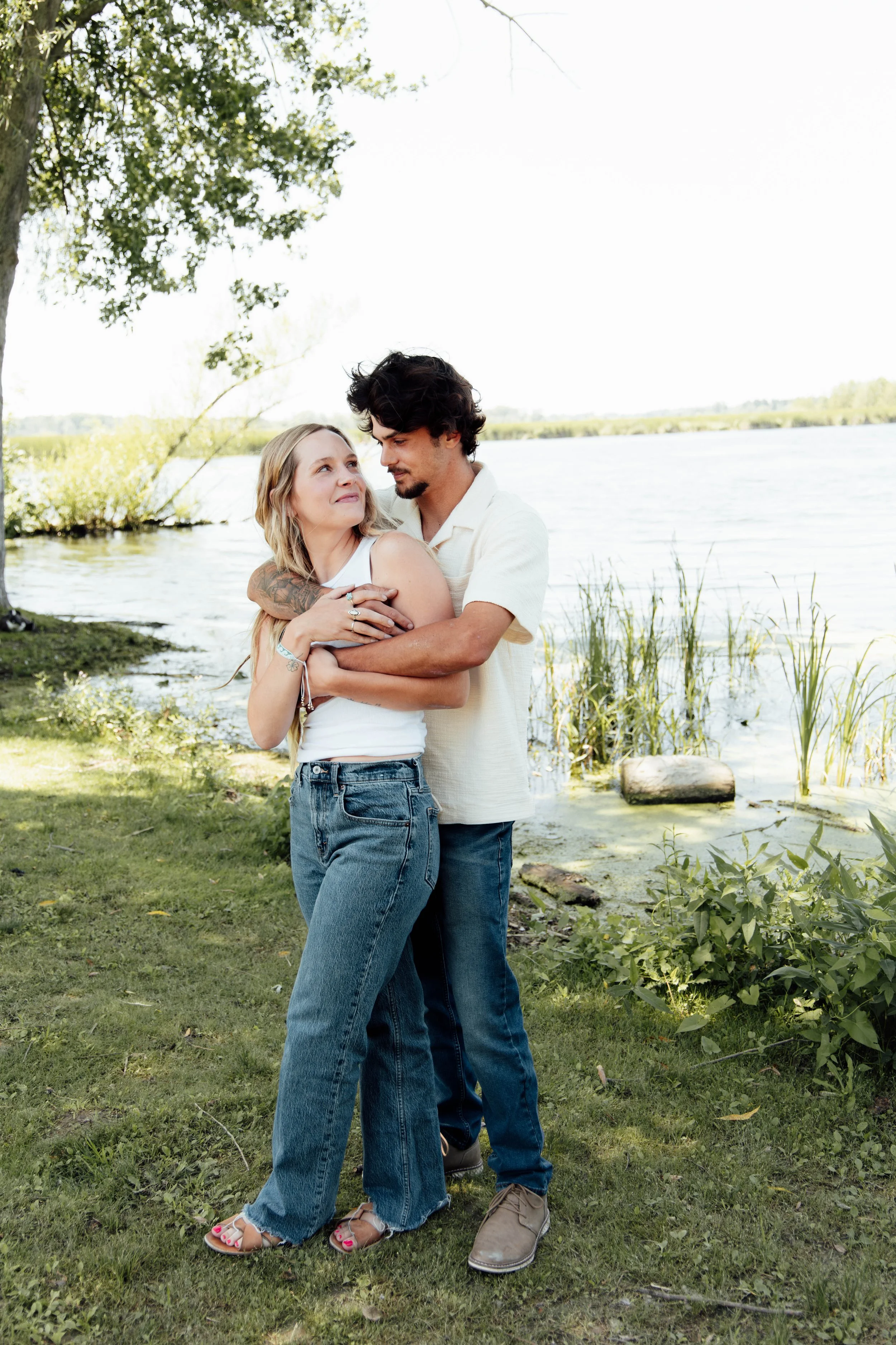 A young woman and man standing together near a lake, embracing each other with smiles, surrounded by trees and grass.