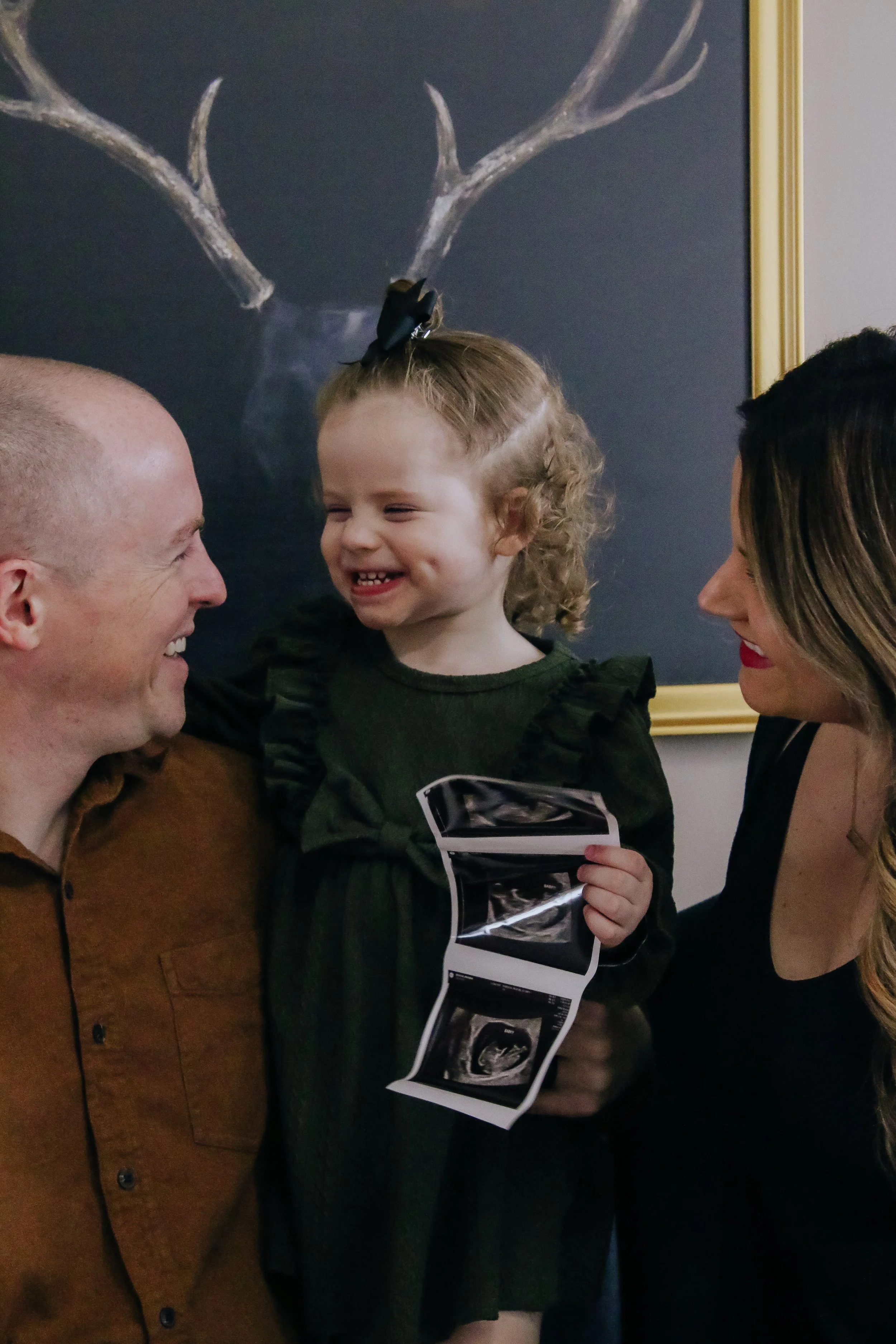A happy family celebrating, with a young girl holding ultrasound pictures, a man and a woman smiling at her, in front of a chalkboard with a reindeer drawing.