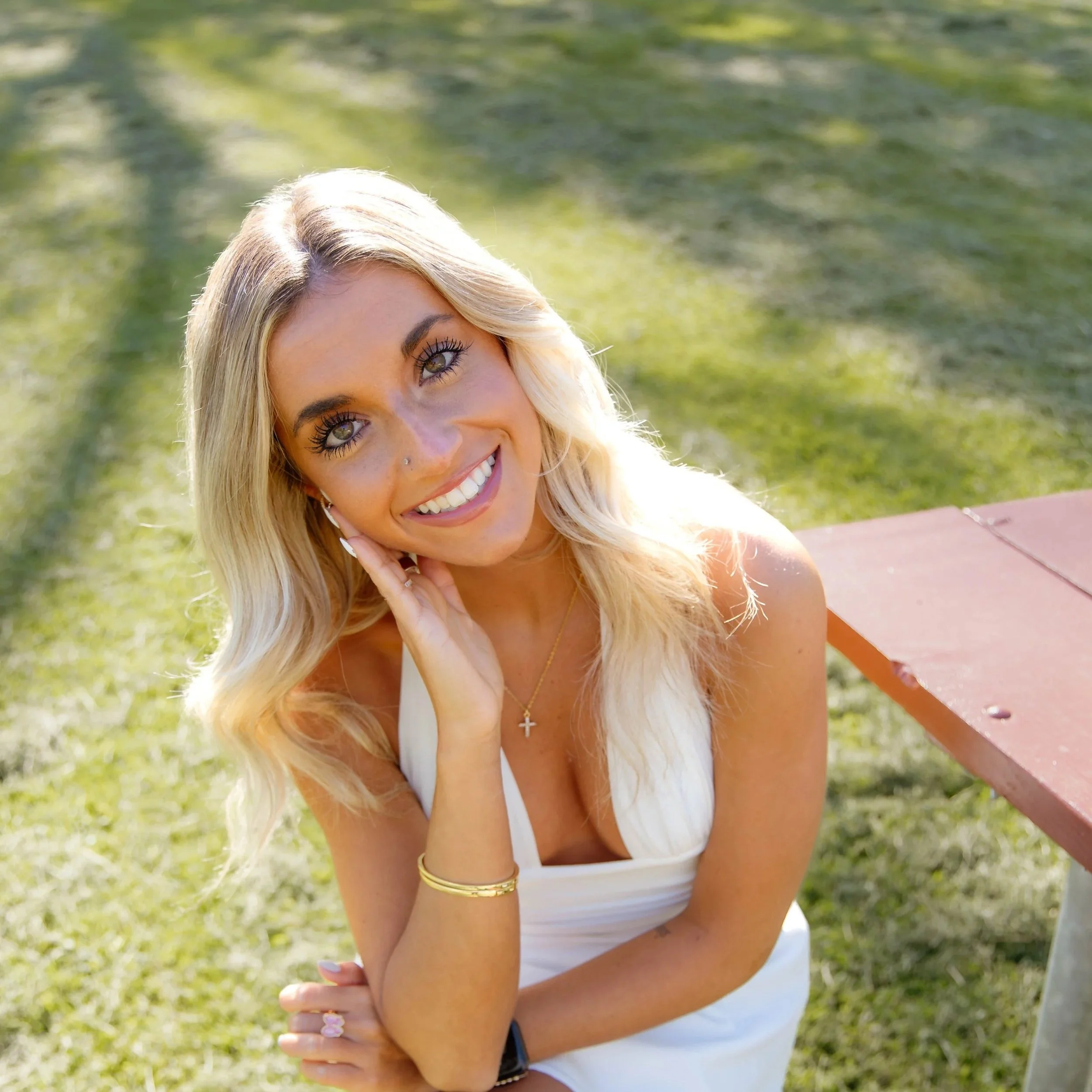 Smiling woman with blonde hair, blue eyes, wearing a white top, gold jewelry, sitting outdoors near a body of water with greenery in the background.