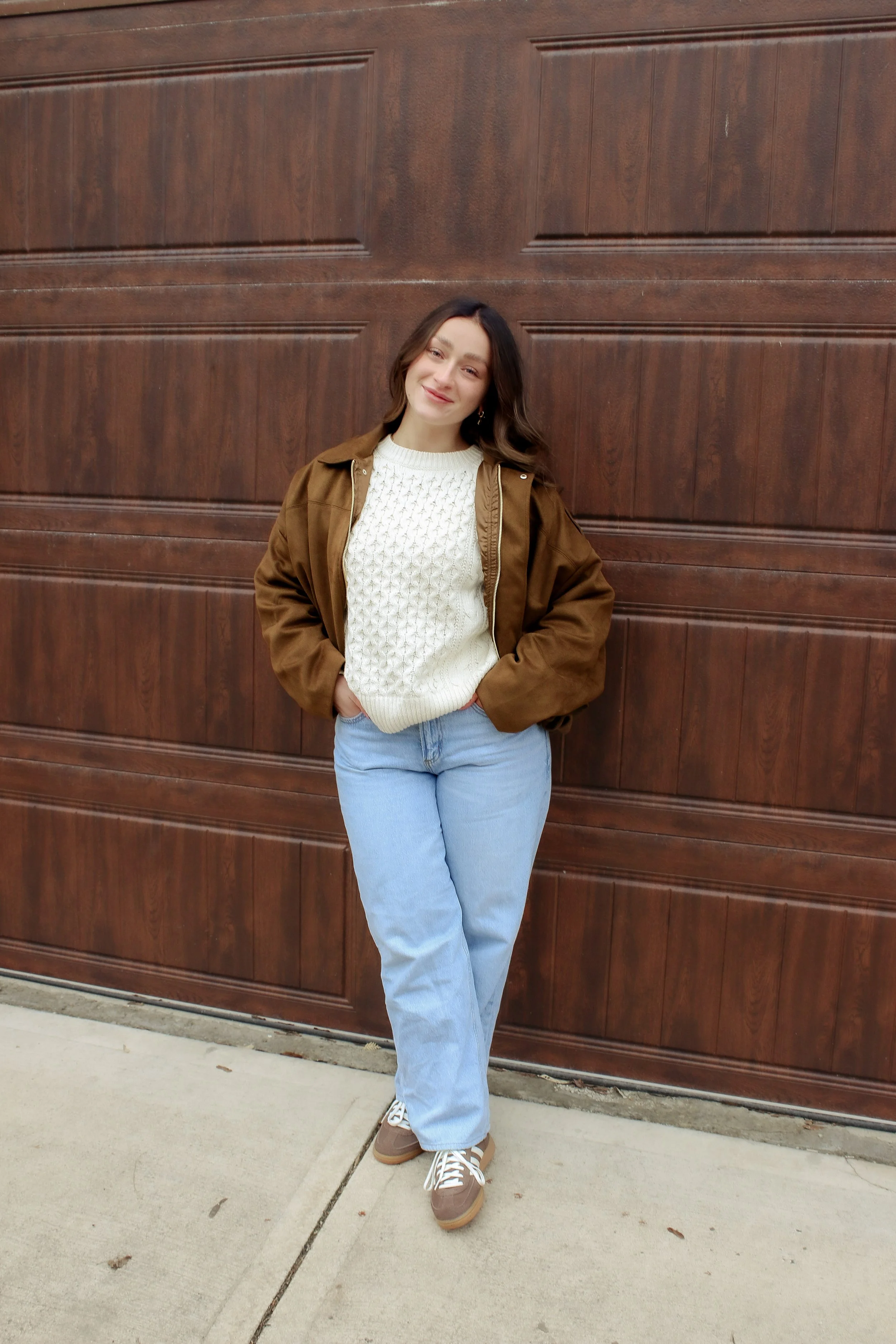 A woman standing outdoors in front of a wooden garage door, wearing a brown jacket, white sweater, light blue jeans, and sneakers, smiling with hands in pockets.
