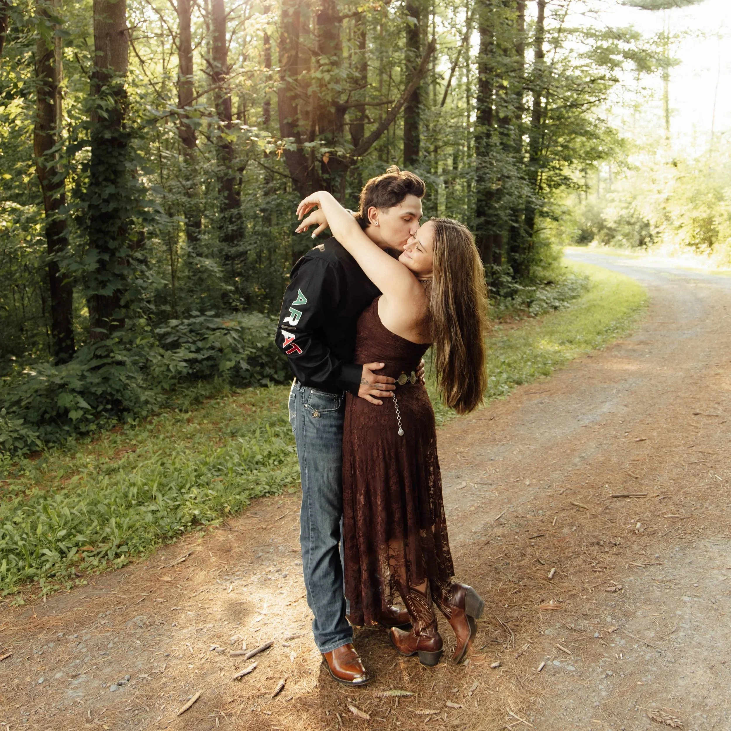 A young man and woman embracing and kissing in a wooded area during daylight. The man is wearing a black jacket with colorful patches, and the woman is dressed in a brown lace dress and cowboy boots. They are standing on a dirt path surrounded by trees and greenery.