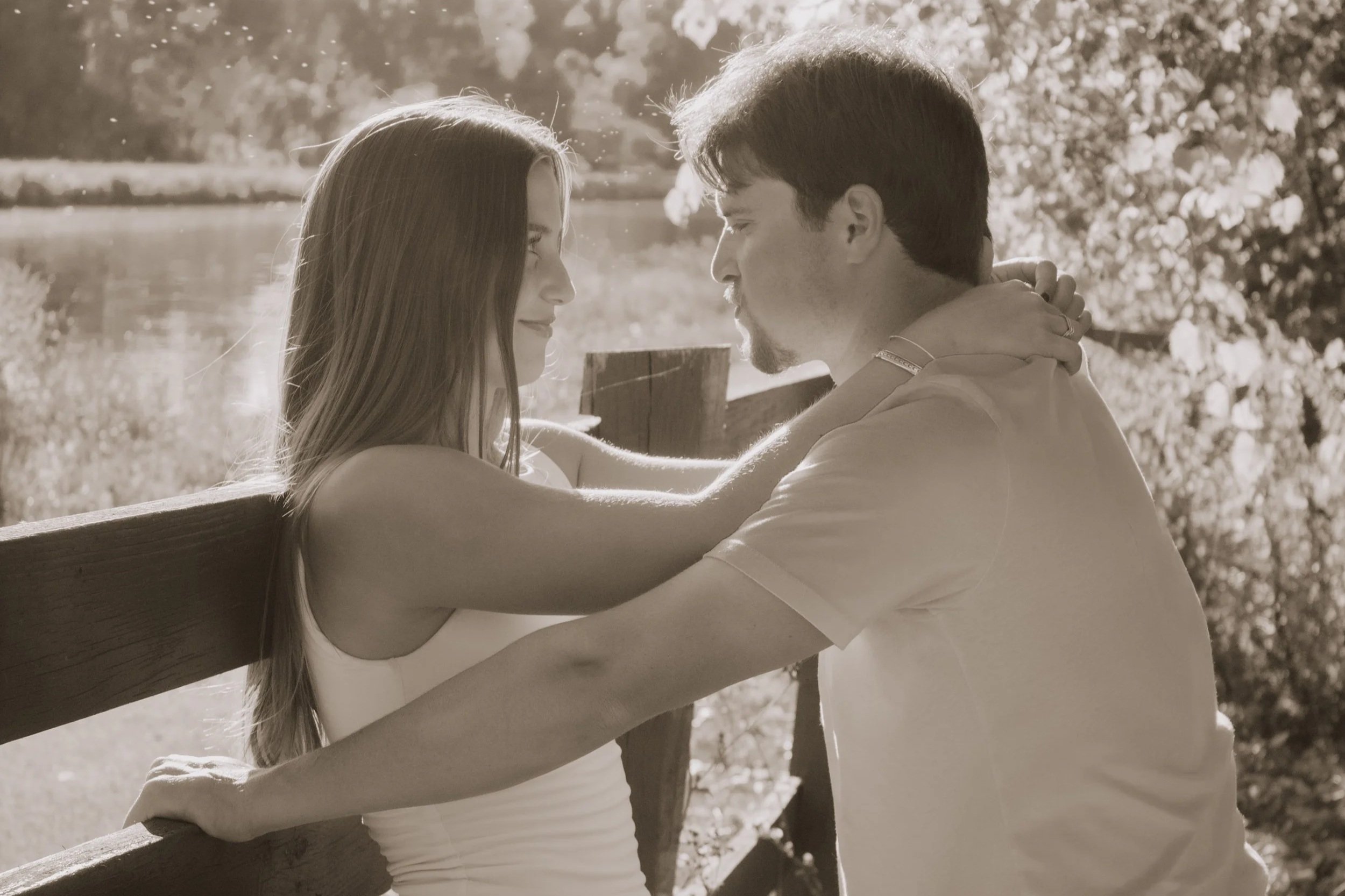 A sepia-toned photograph of a couple embracing outdoors near a wooden railing, with trees and a body of water in the background, sharing a tender moment.